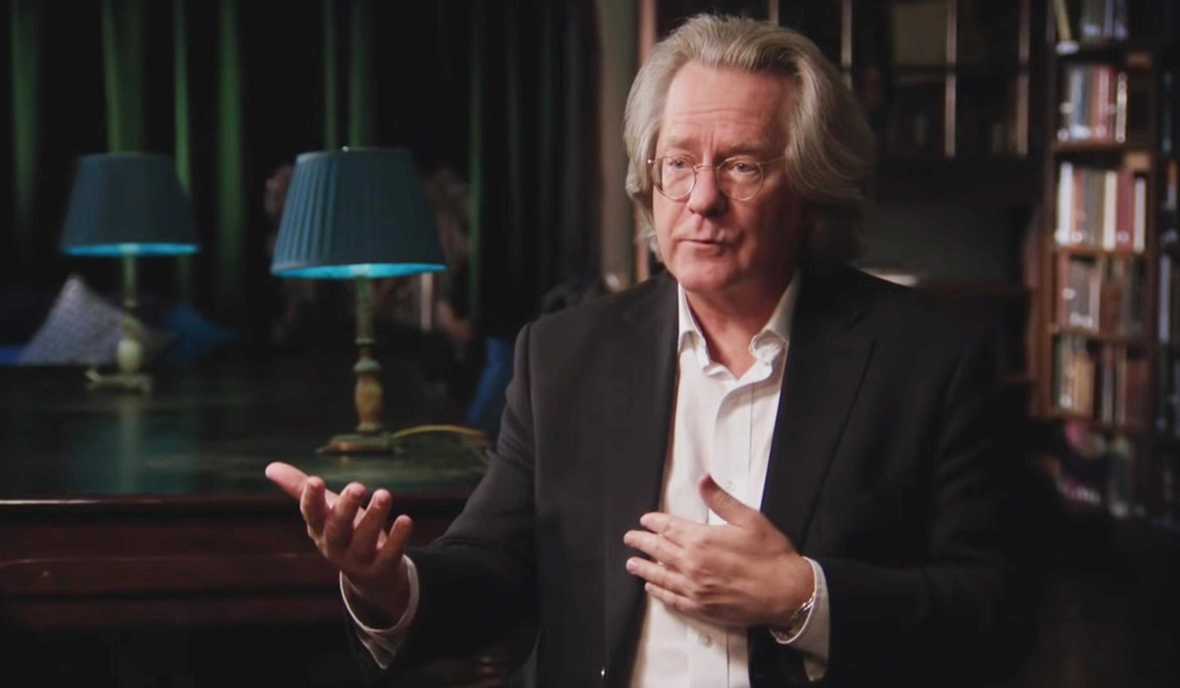 Man with glasses and long hair in a dark suit, gesturing while speaking in a dimly lit room with bookshelves and two blue lamps in the background.