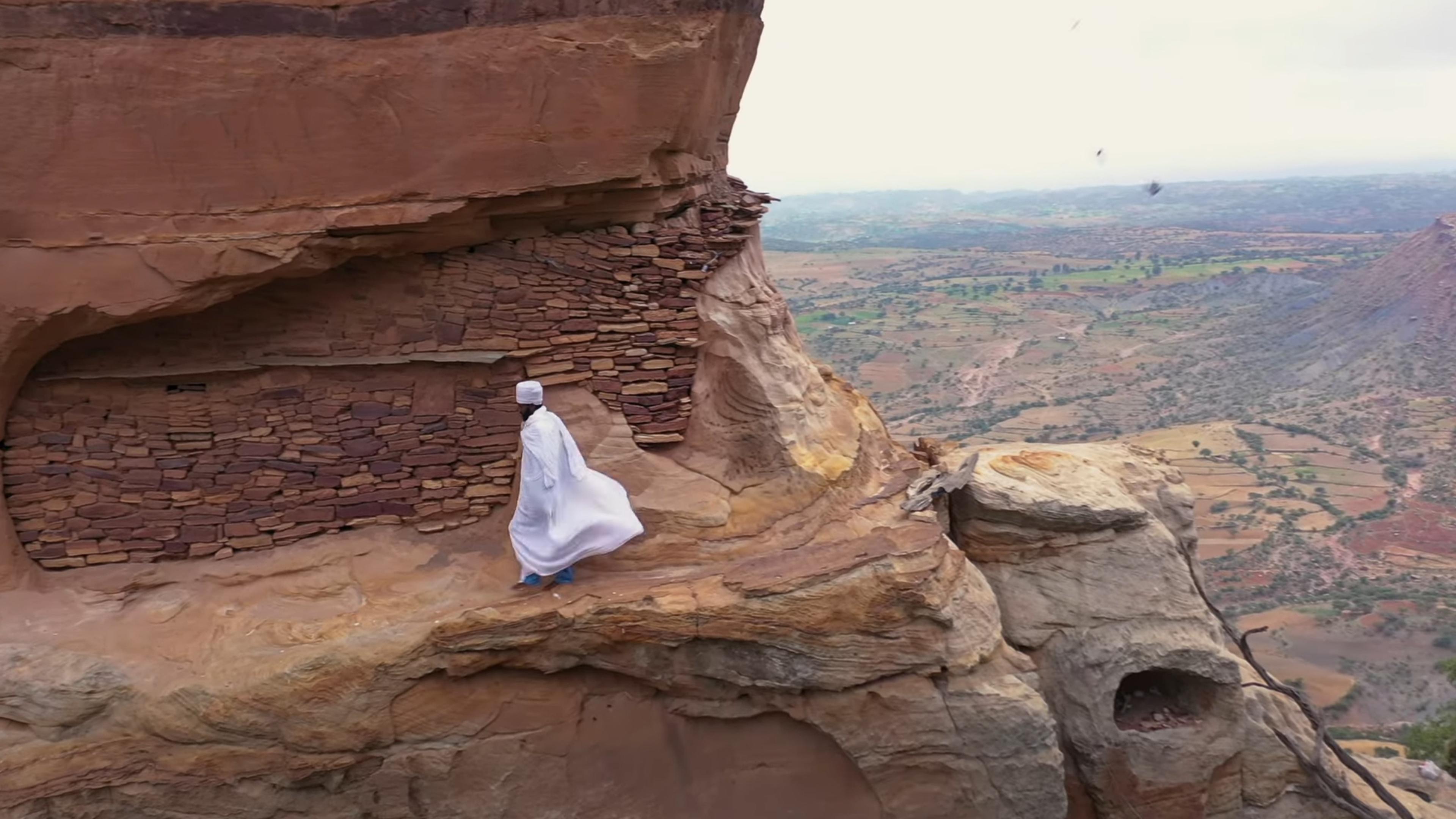 Photo of a person in white walking along a cliffside path near a stone structure with a scenic landscape view.