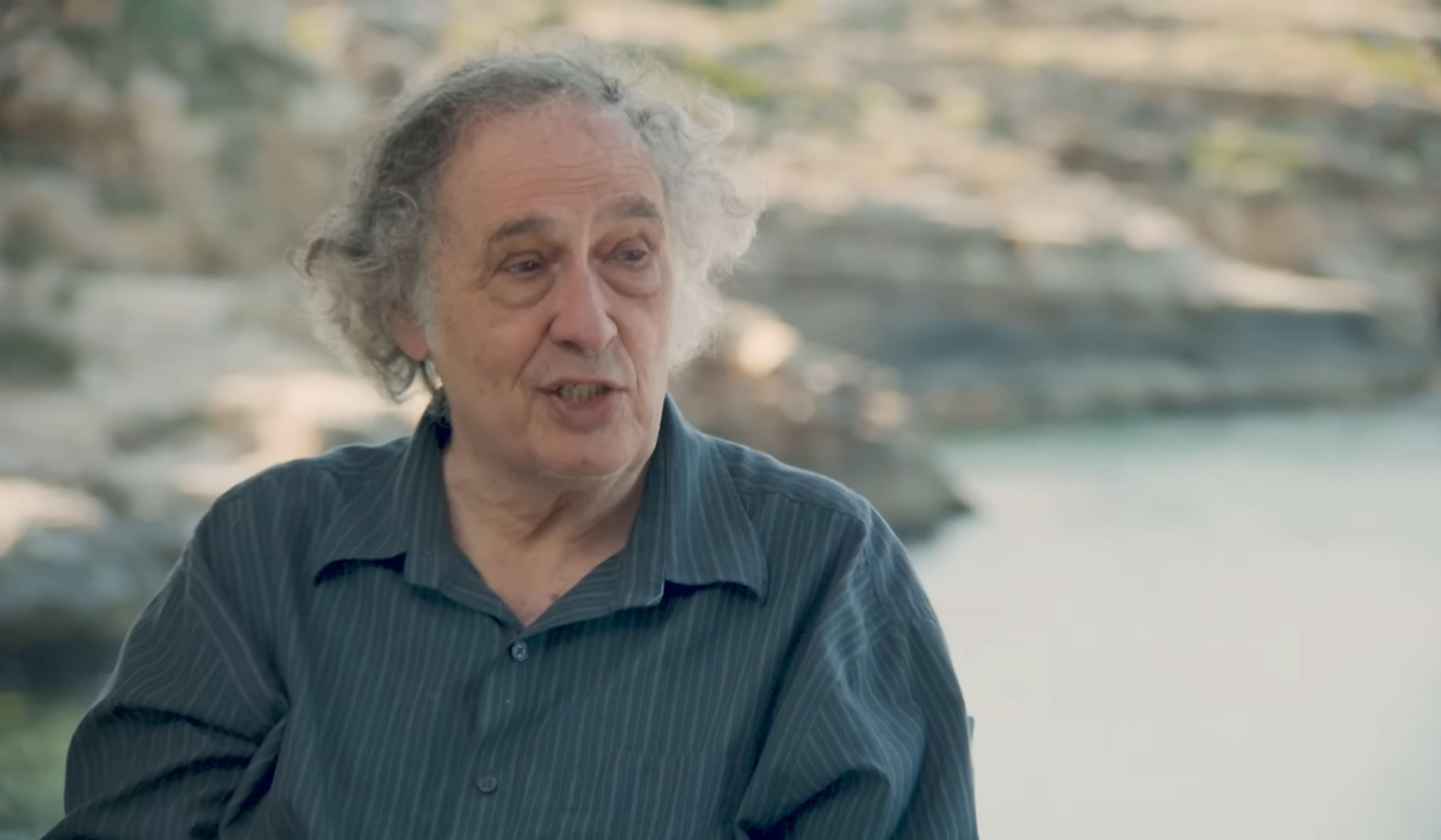 An older man with curly grey hair and a striped shirt speaking outdoors, with a blurred background of rocks and water.