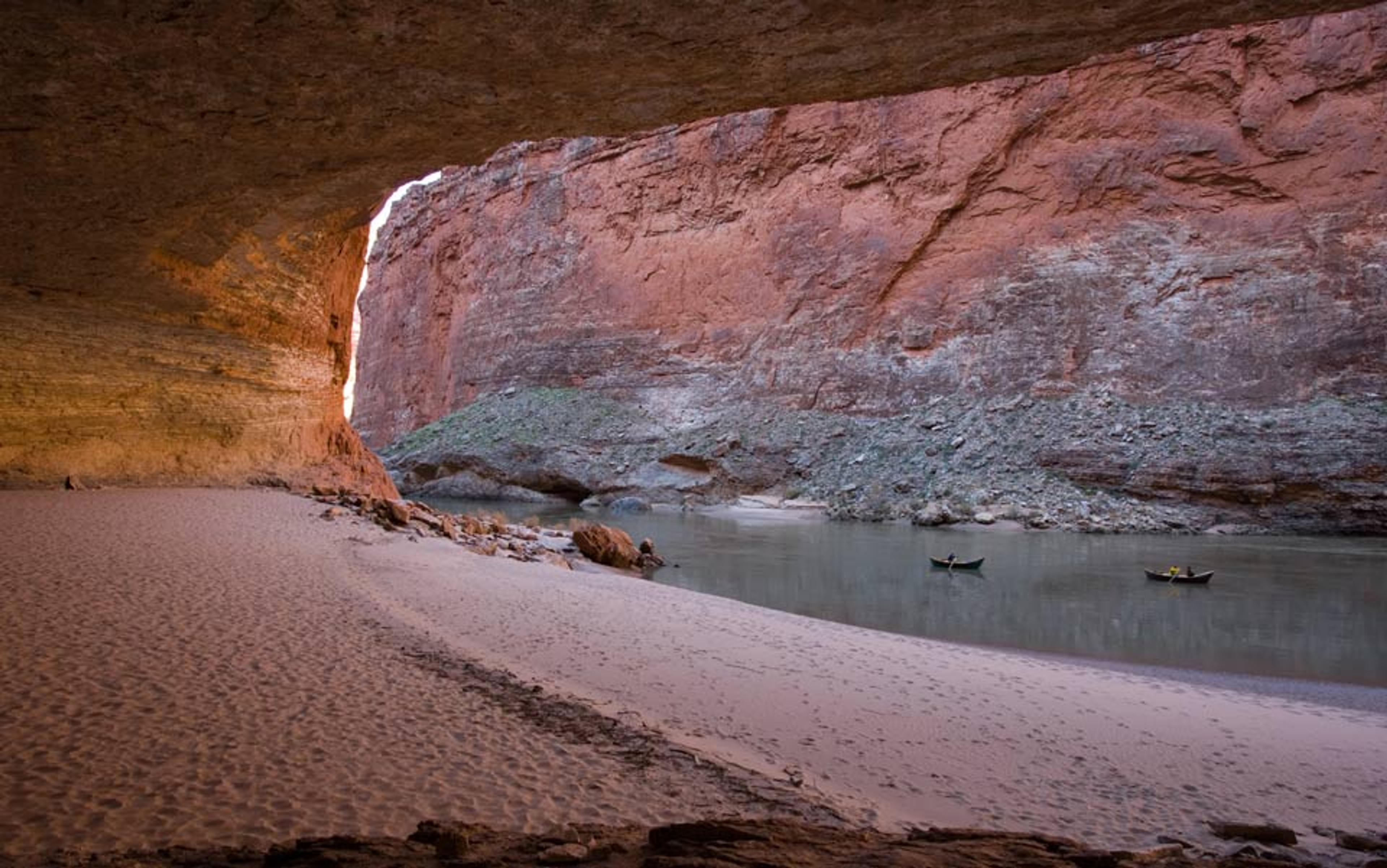 A river with two kayaks, surrounded by steep canyon walls, taken from inside a cave-like area with sandy banks.