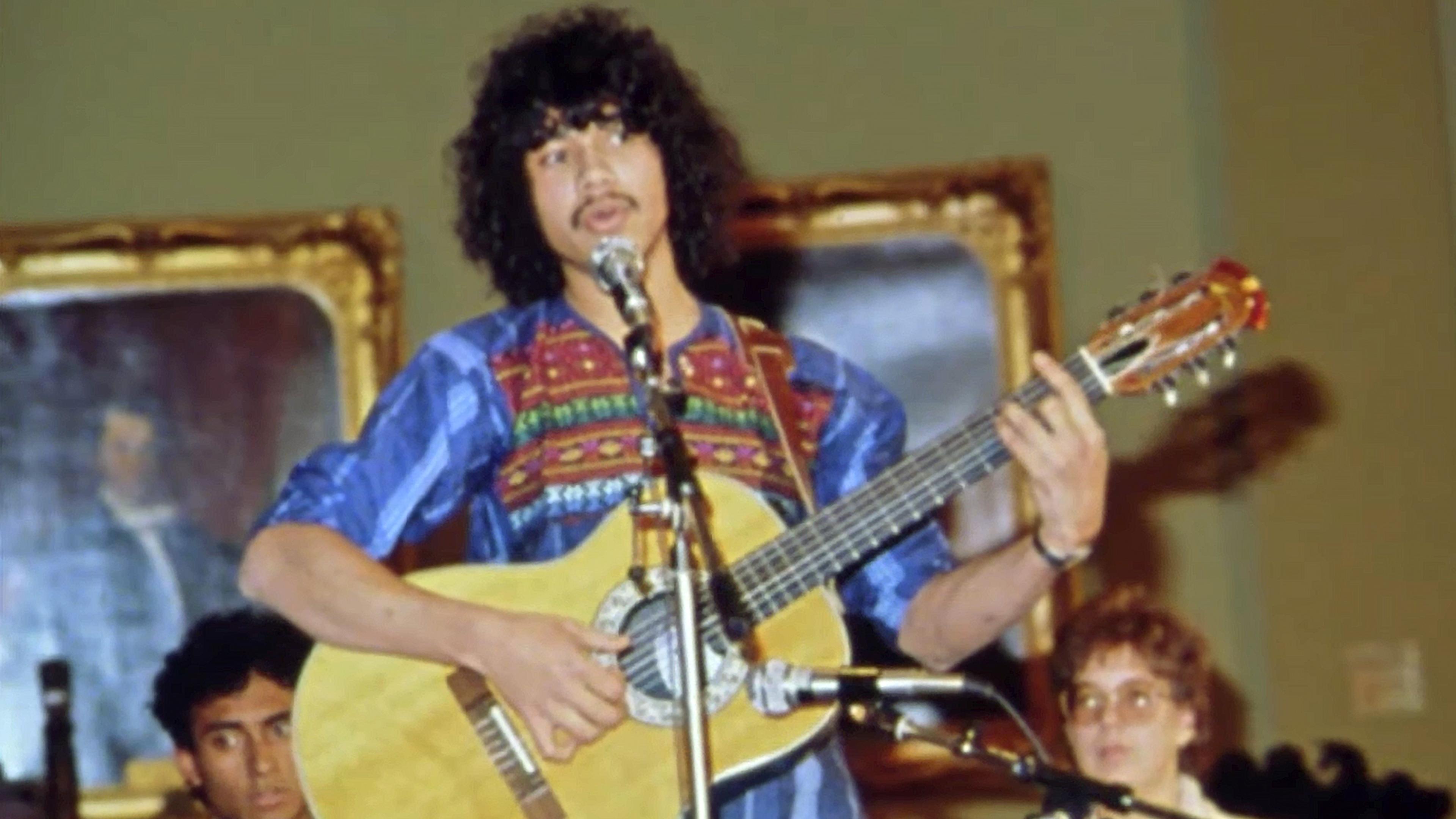 Photo of a musician with curly hair playing acoustic guitar on stage wearing a colourful shirt with audience members in the background.