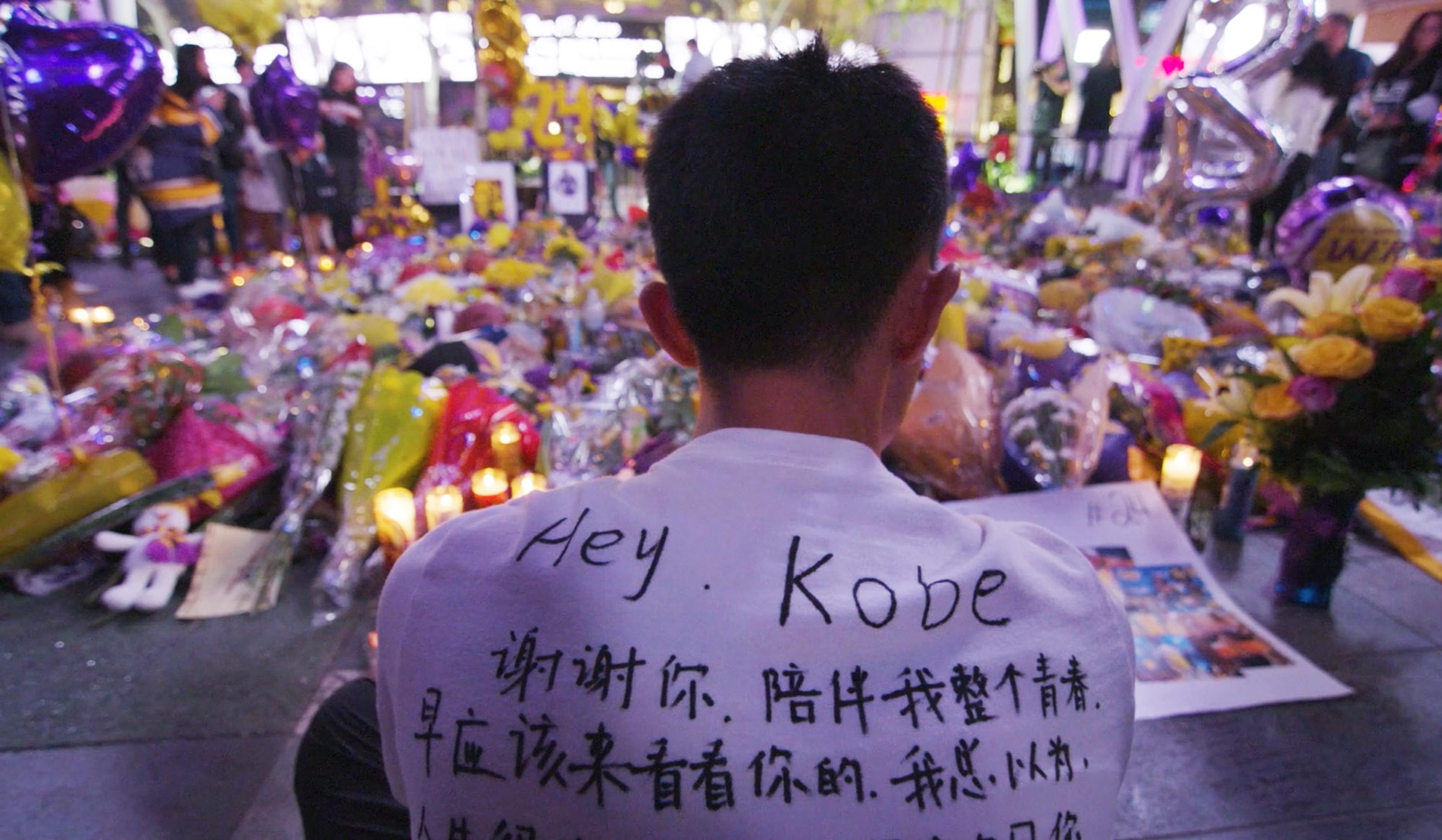 A man in a white shirt with the text ‘Hey Kobe’ and Kanji script sits before a makeshift memorial of flowers, candles, and balloons.