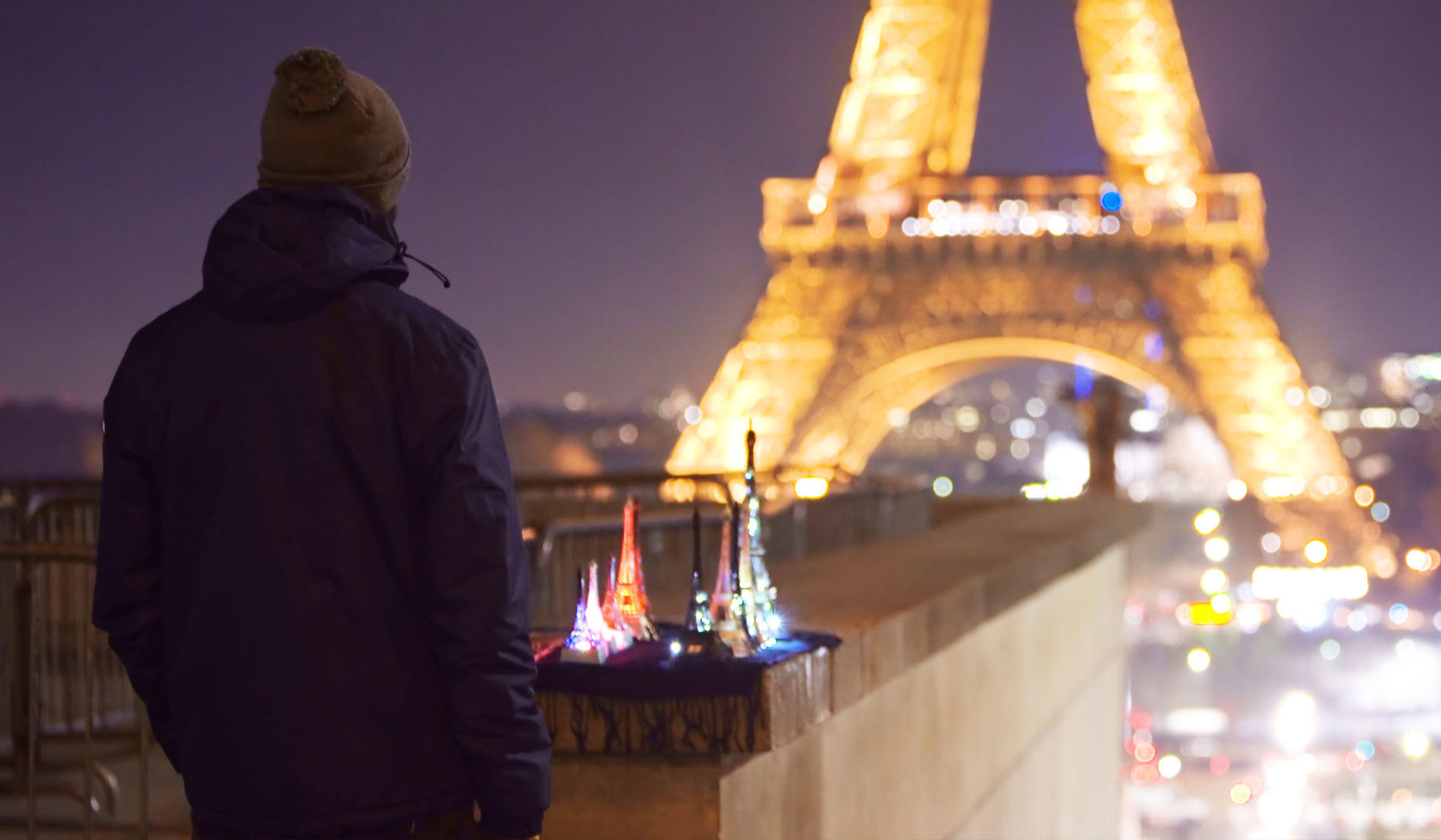 A person in a jacket and hat stands at night near illuminated Eiffel Tower models with the actual Eiffel Tower in the background.