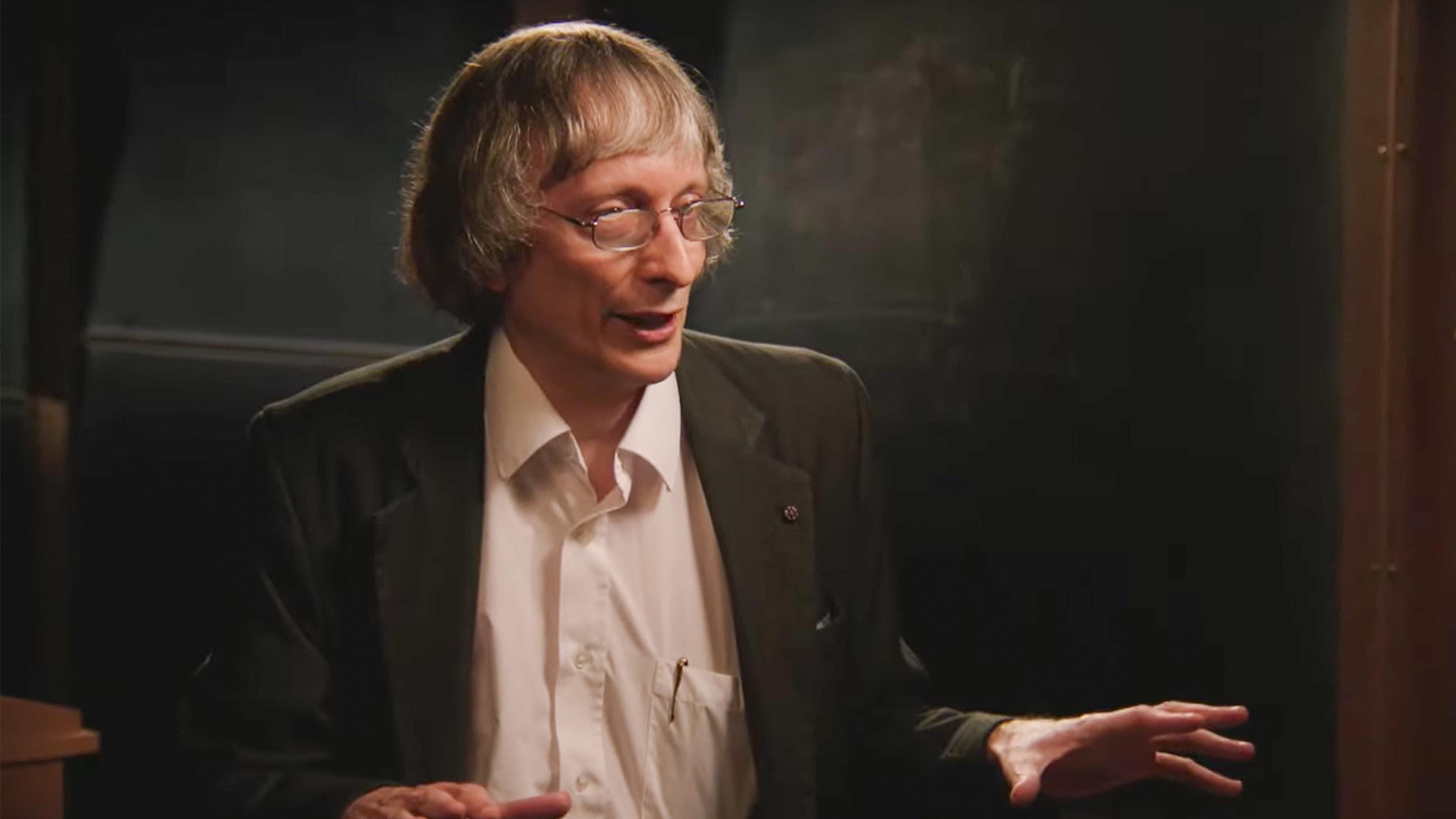 Photo of a man with glasses and grey hair in a suit speaking animatedly against a dark background.