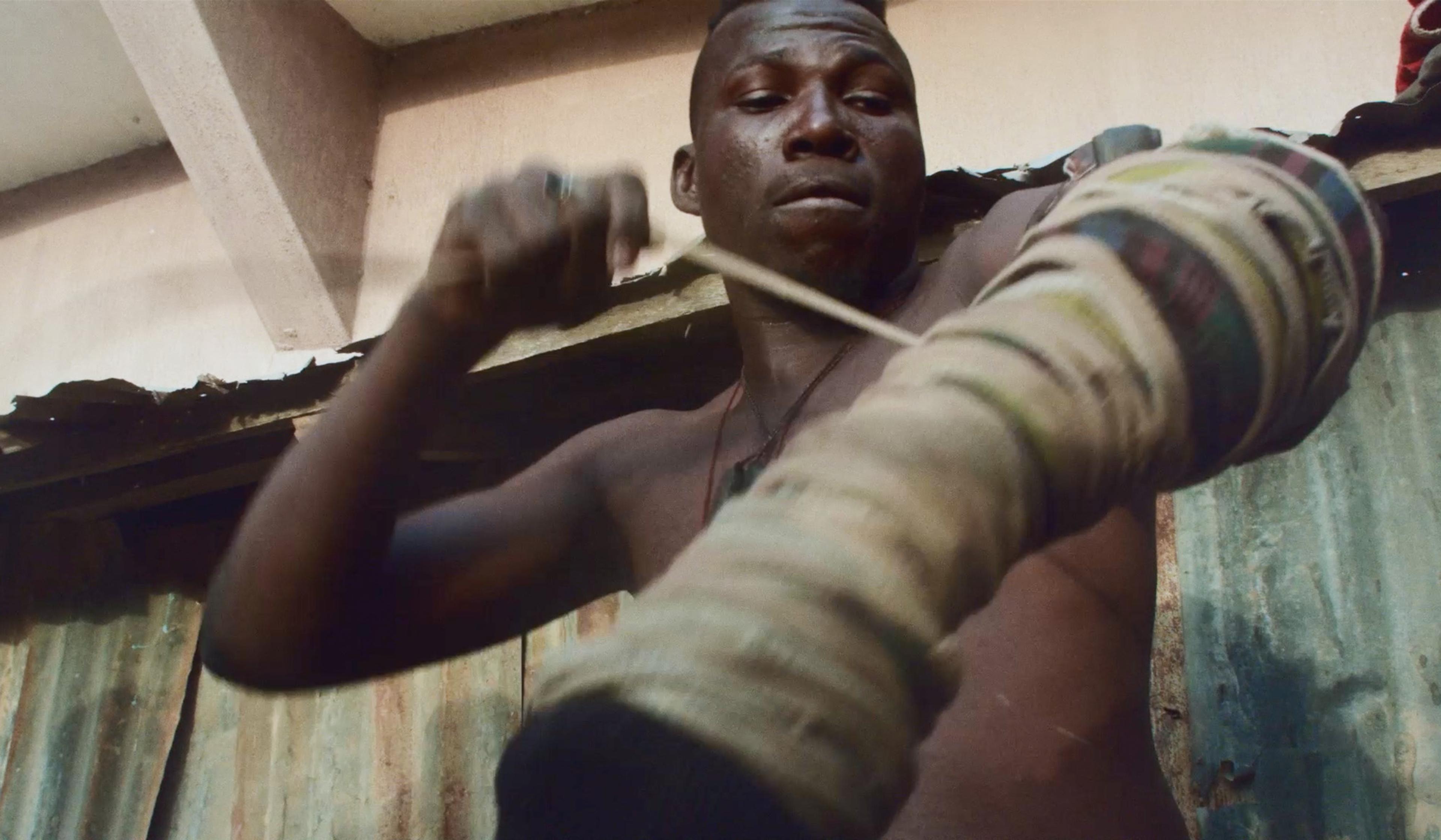 A man in a rural setting wraps his hands and prepares for physical work or training, with a focused expression and a rustic tin roof in the background.