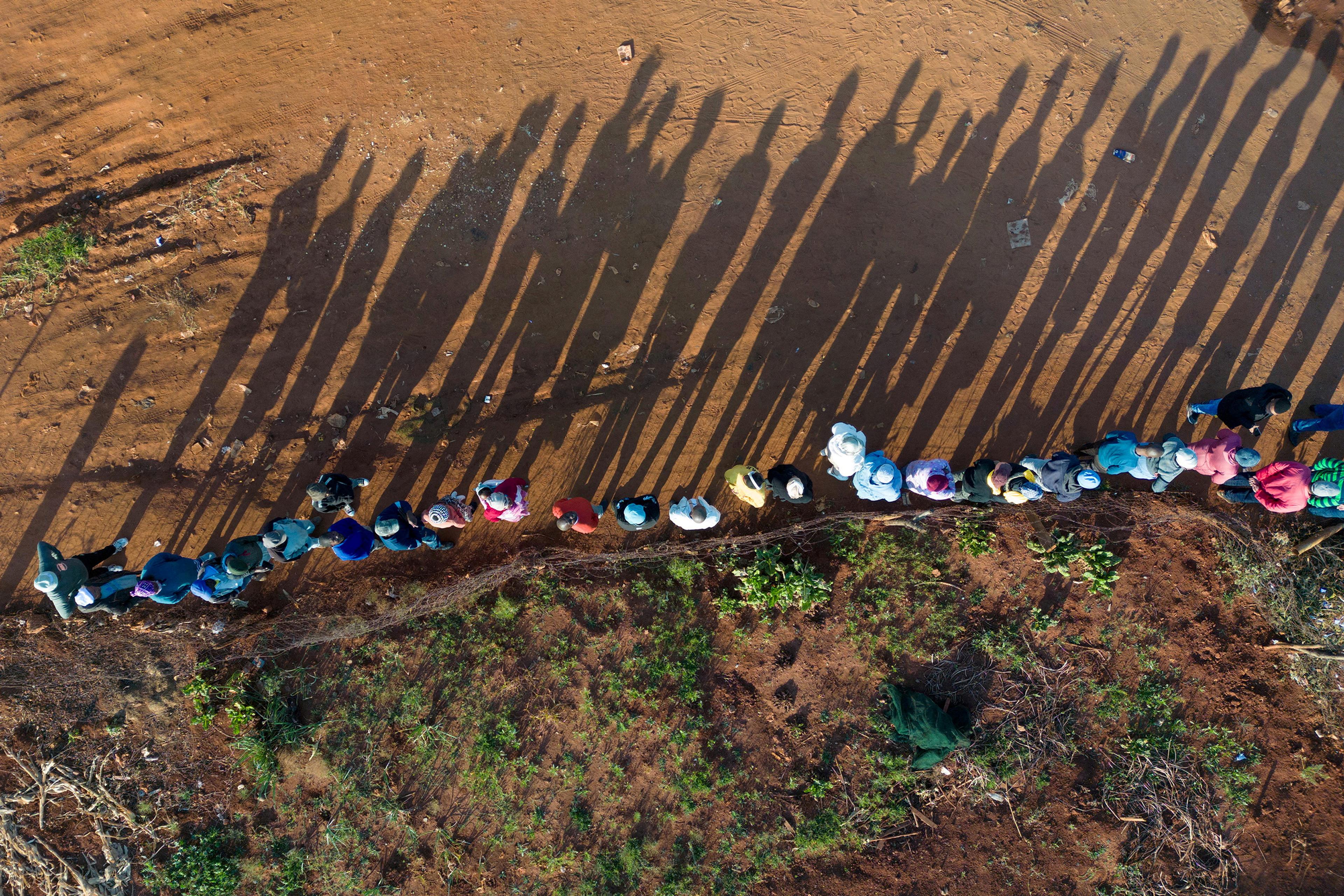 Aerial photo of people in colourful clothes standing in line casting long shadows on a dirt path surrounded by greenery.
