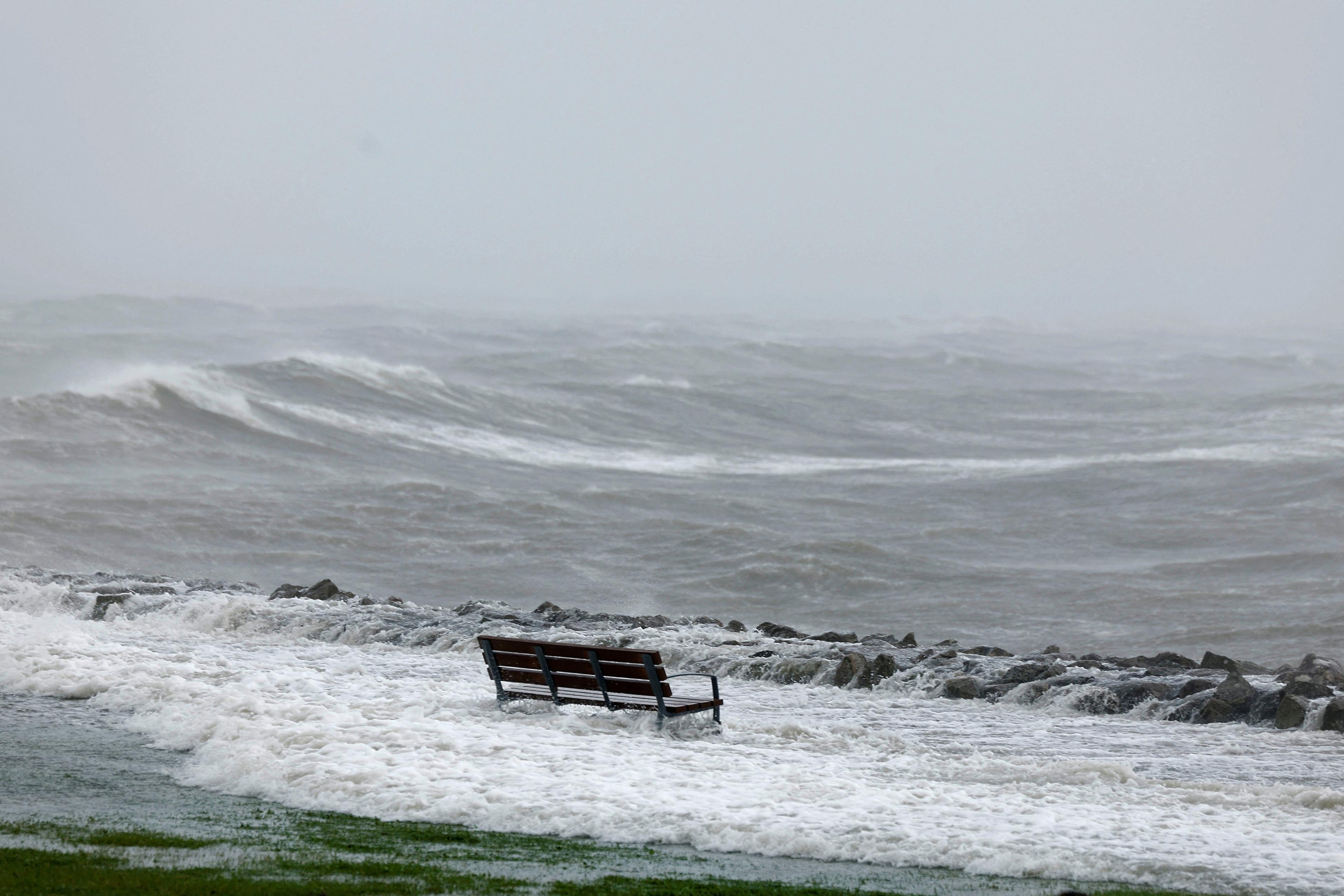 Photo of a lone bench facing turbulent ocean waves under a cloudy sky. The area is partially flooded by the sea.