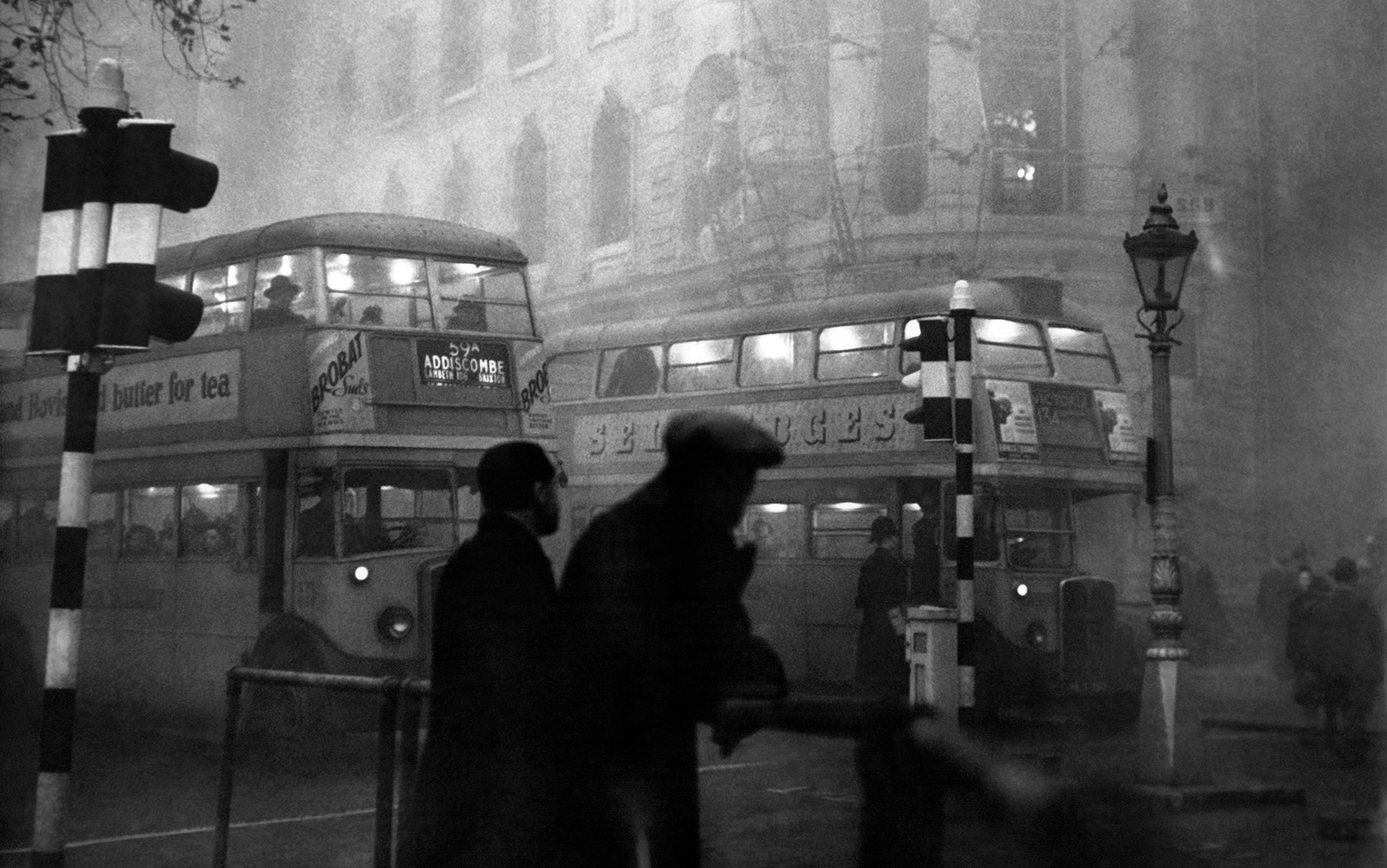 Vintage photograph of two double-decker buses in a foggy city scene with pedestrians and traffic lights in the foreground.