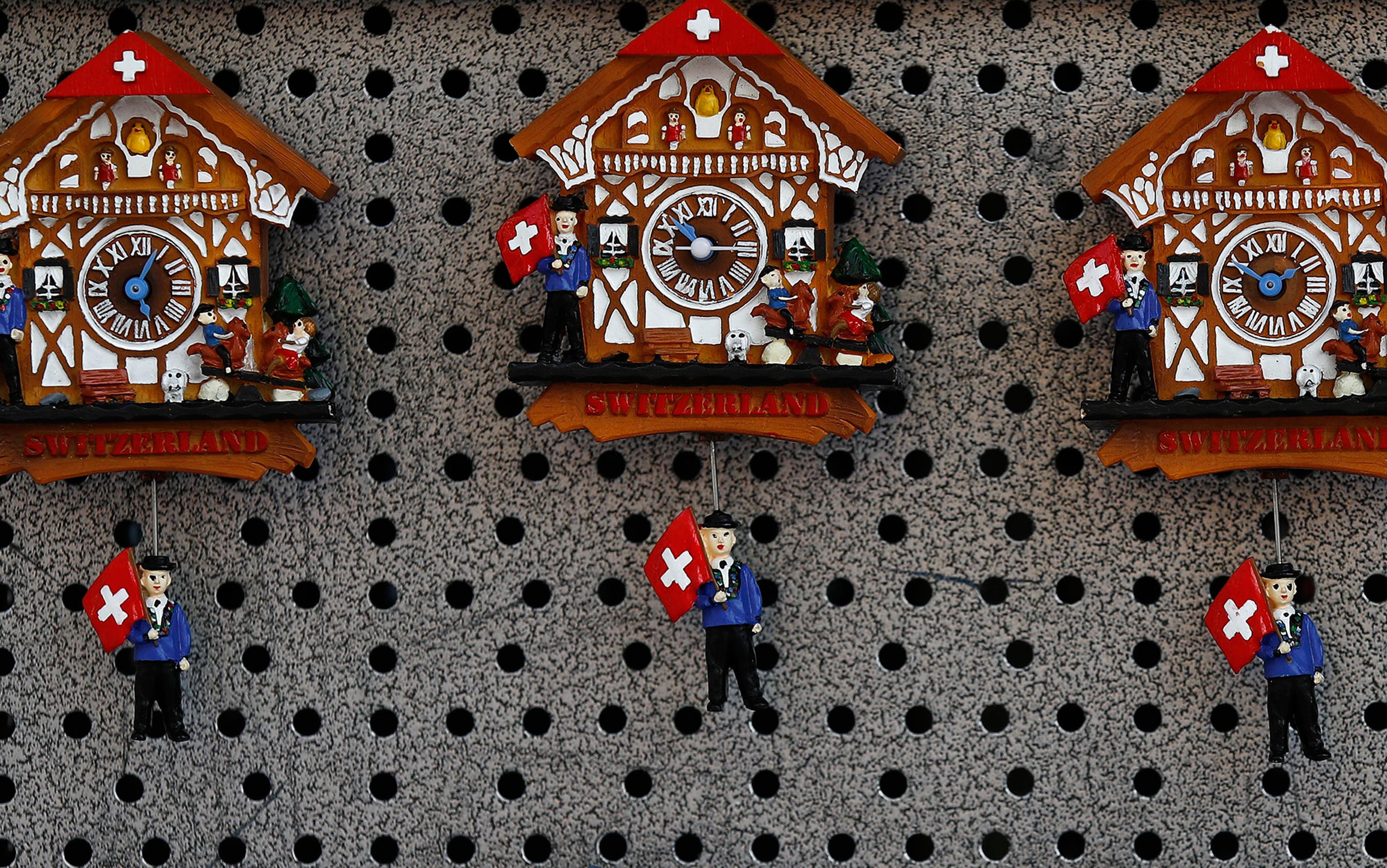 Three cuckoo clocks in the style of Swiss chalets with figurines holding Swiss flags hanging on a perforated display board.