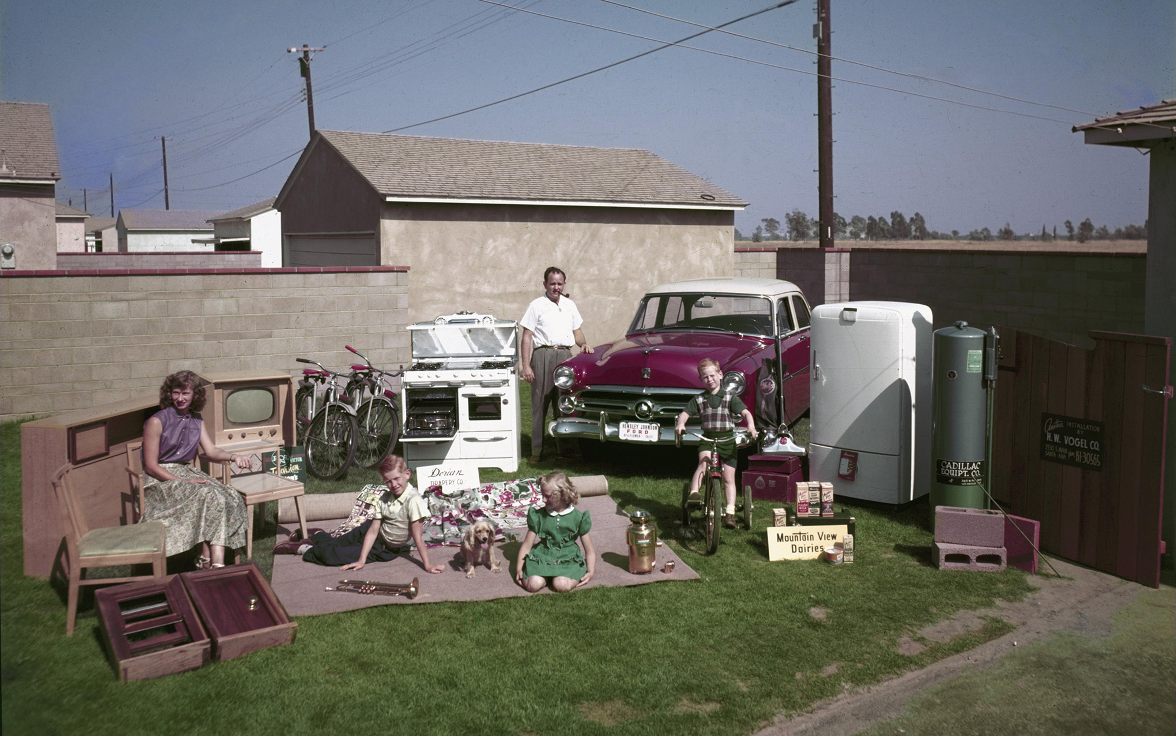 A 1950s family photo with a car, household appliances and children with bicycles and toys in a suburban backyard setting.