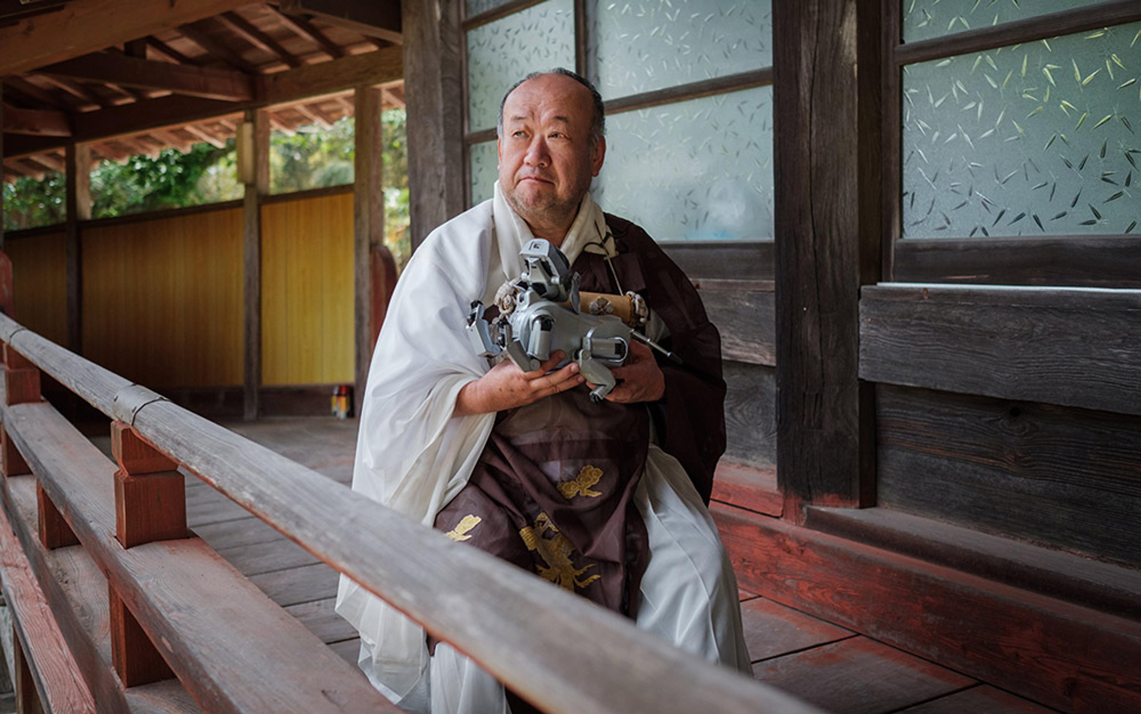 A Japanese man in traditional attire holding a grey robotic arm, seated on a wooden porch, next to glass-paned doors and wooden walls.