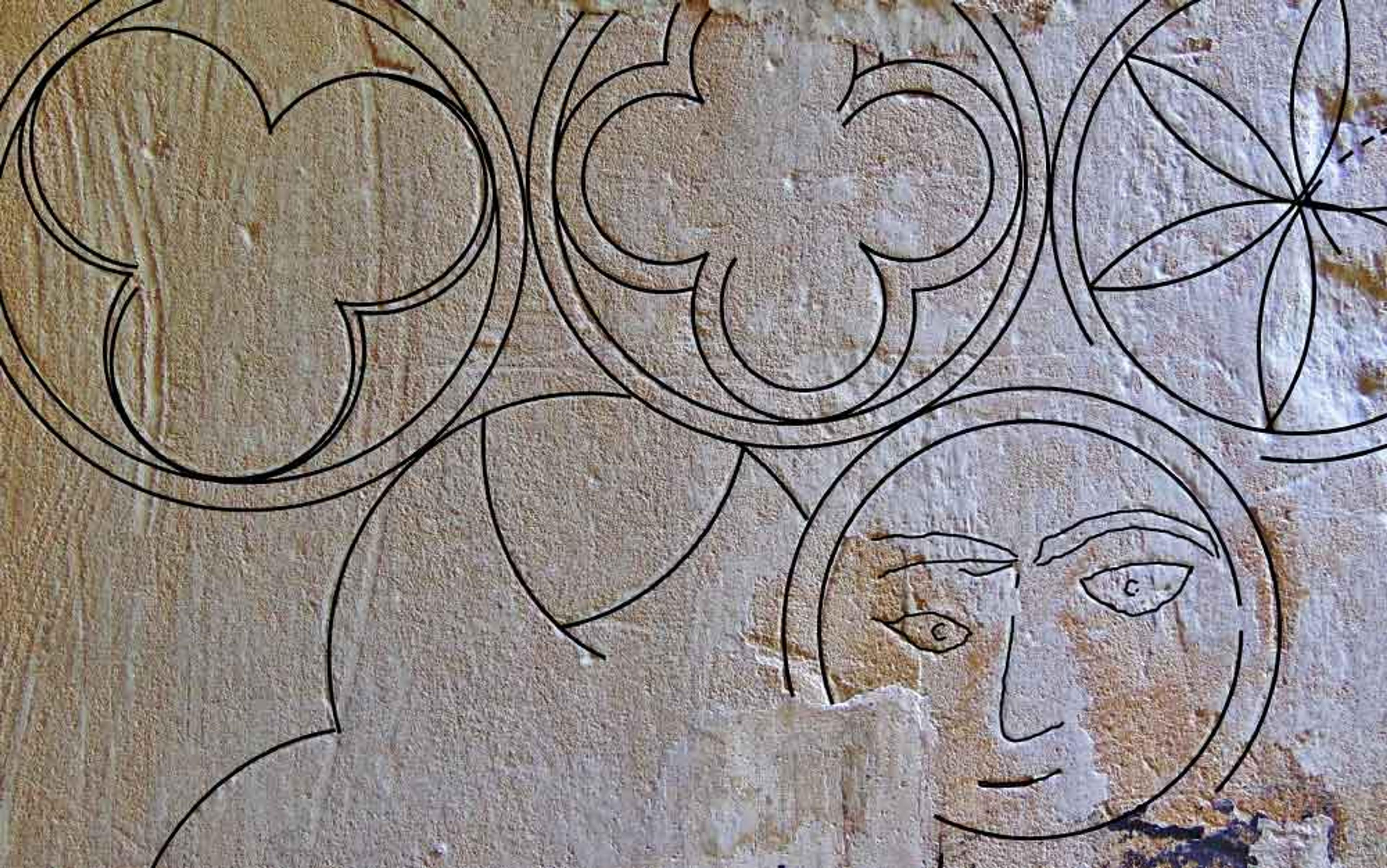 Drawing of geometric shapes and a face carved into a textured stone wall, including circles and flower designs.