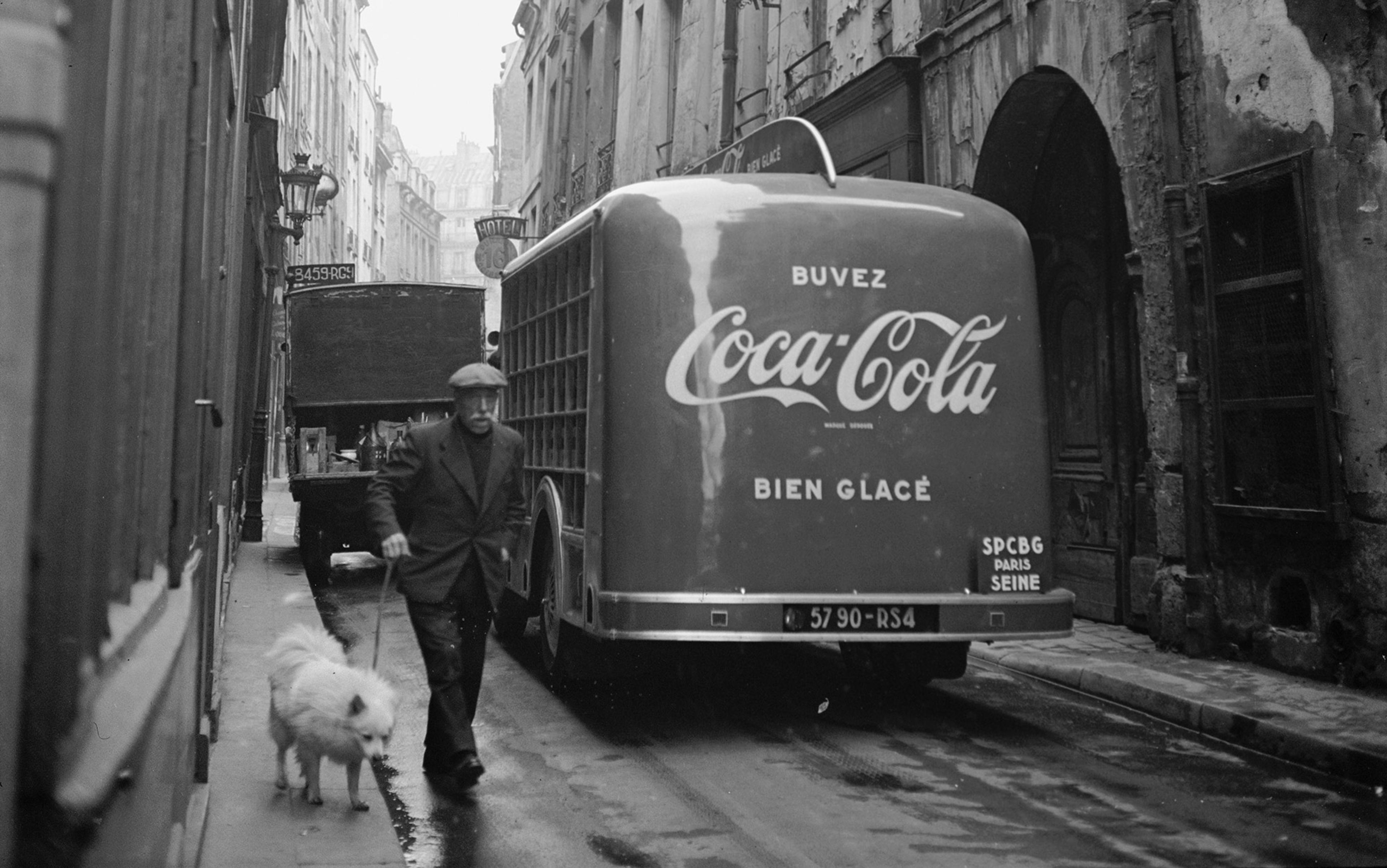 Black and white photo of a man walking a dog in a narrow street, with a Coca-Cola delivery truck and old buildings in the background.