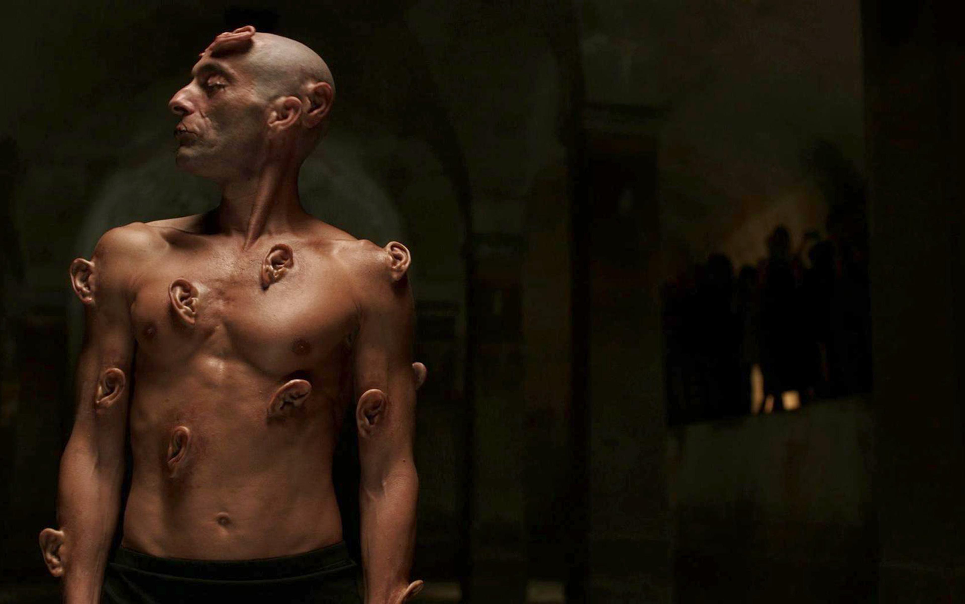 A shirtless man with multiple ears growing out of his body in strange places, in a dimly lit room.
