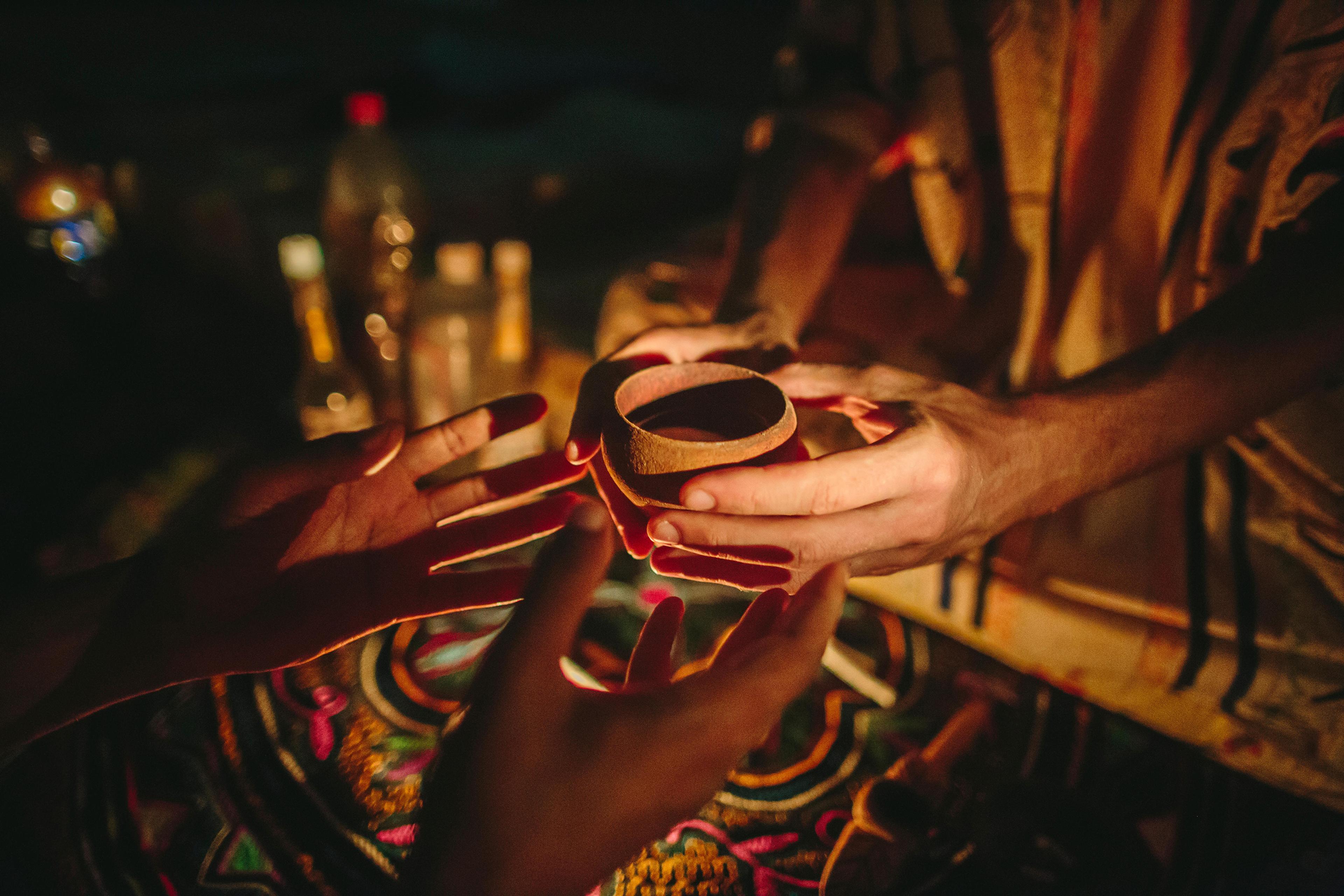 Close-up photo of hands passing a small wooden cup during a dimly lit gathering with bottles visible in the background.