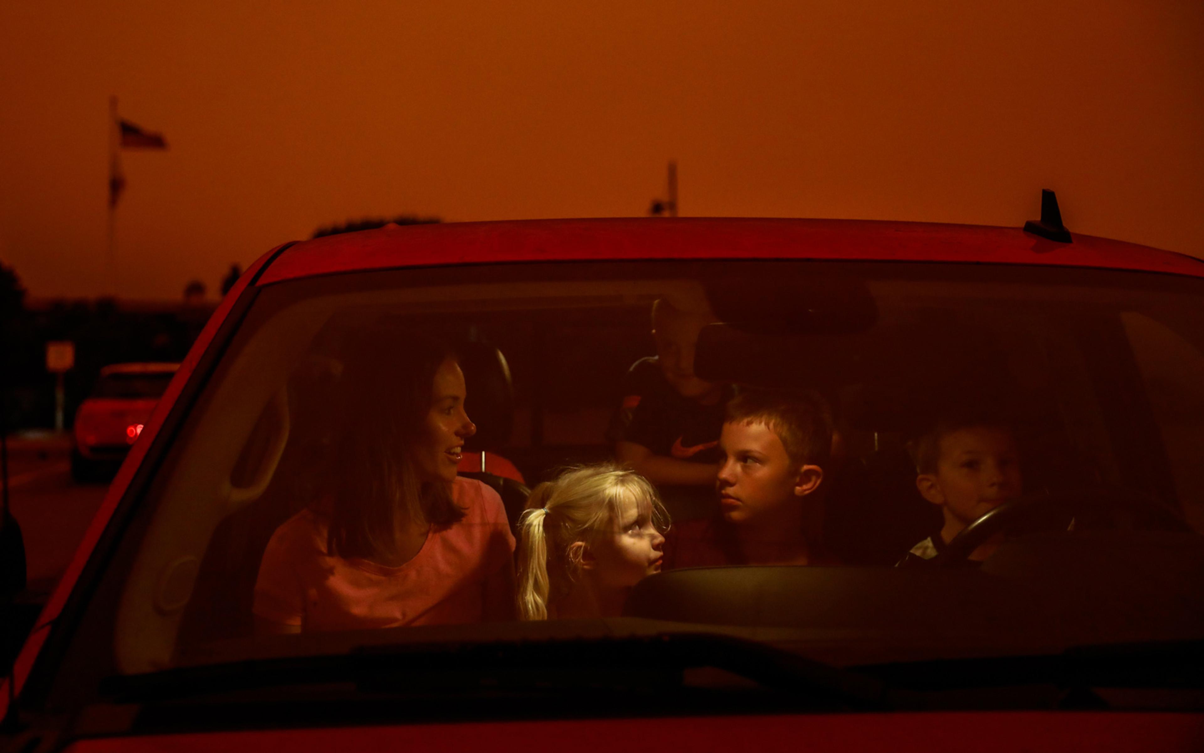 A woman and four children inside a car, under an orange sky. A boy is in the driver’s seat, the woman in the passenger seat.