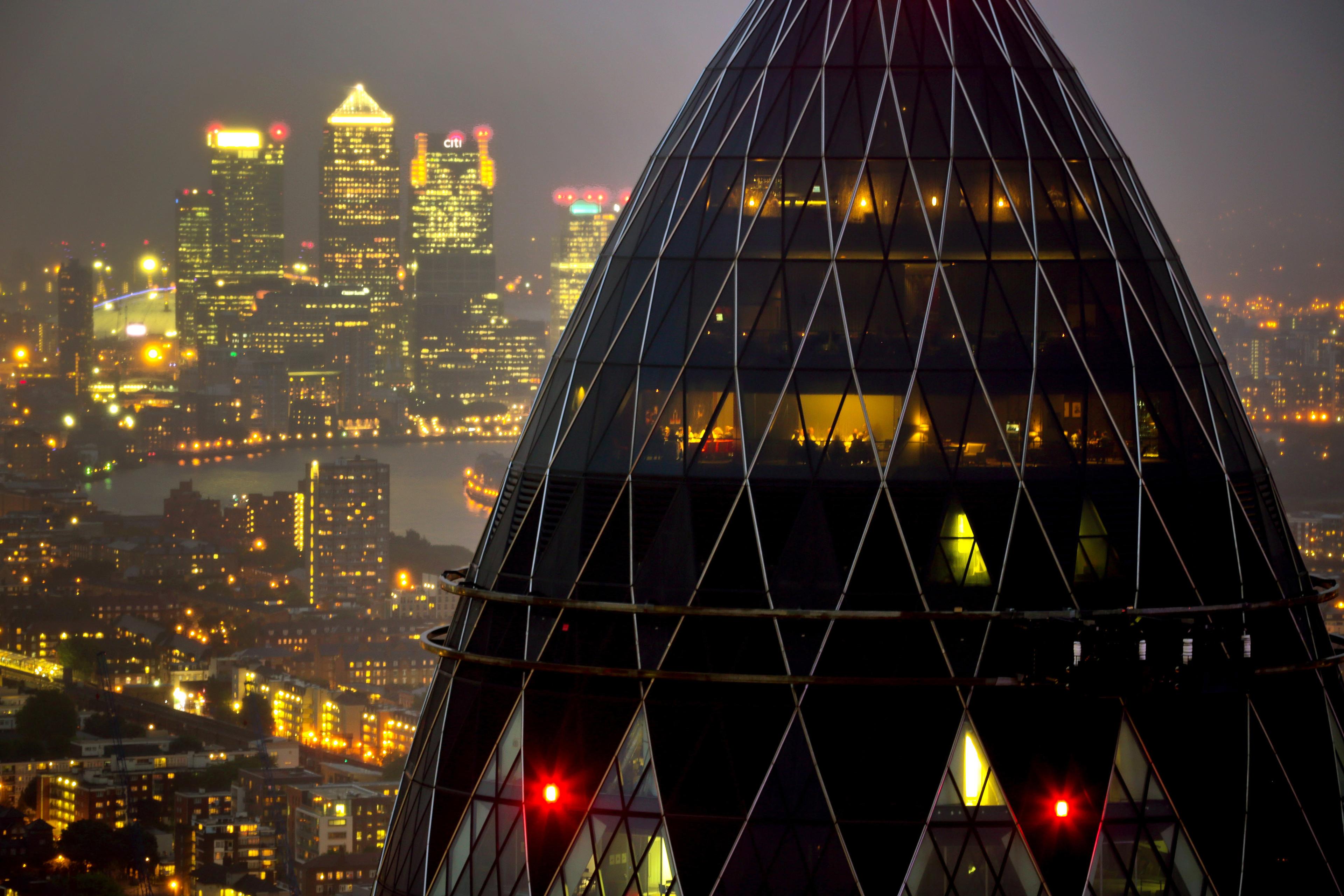 London’s Gherkin building lit at night with Canary Wharf skyscrapers glowing in the hazy background.
