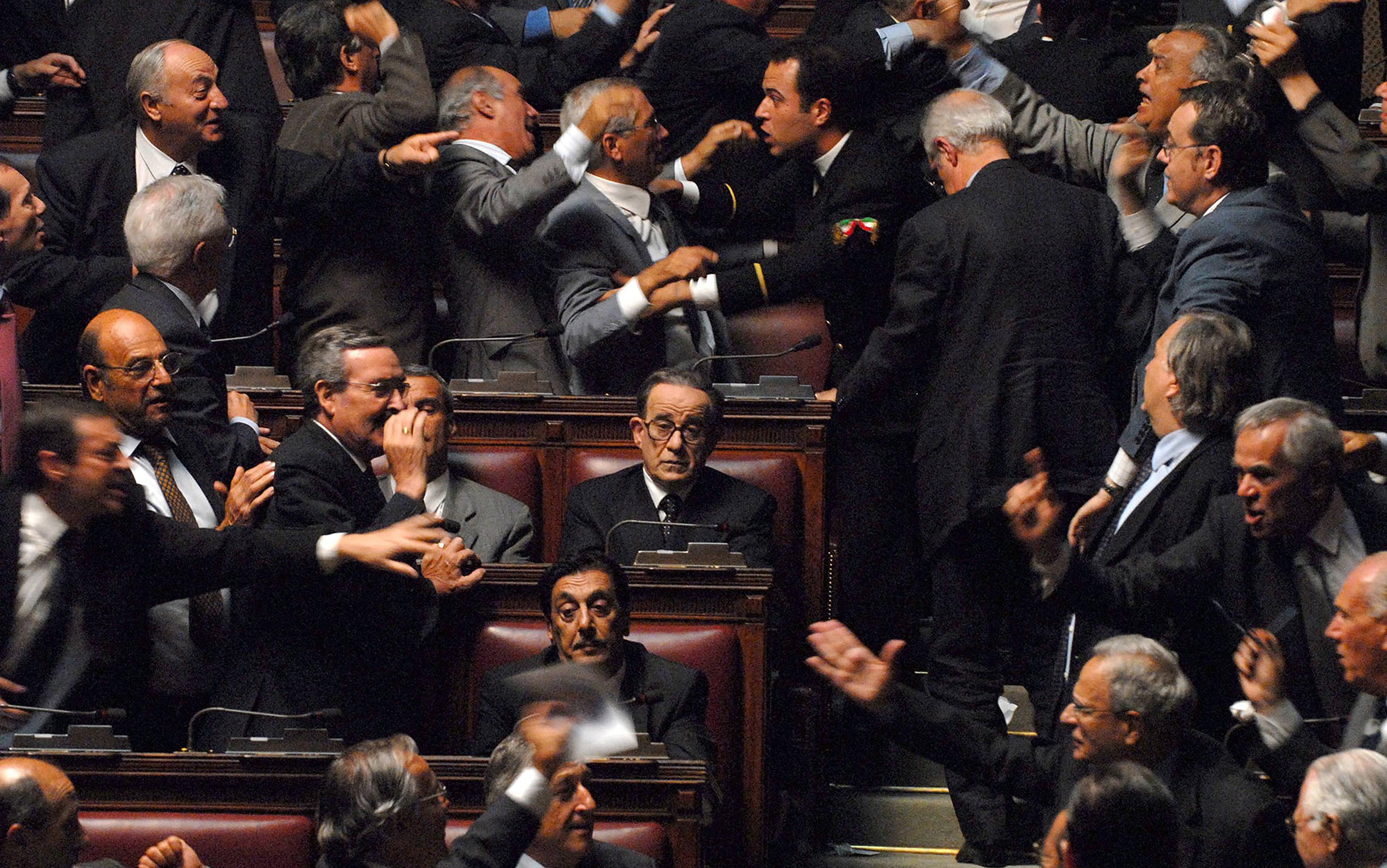 A chaotic parliamentary session with politicians arguing, gesturing and one calm individual seated in the centre.