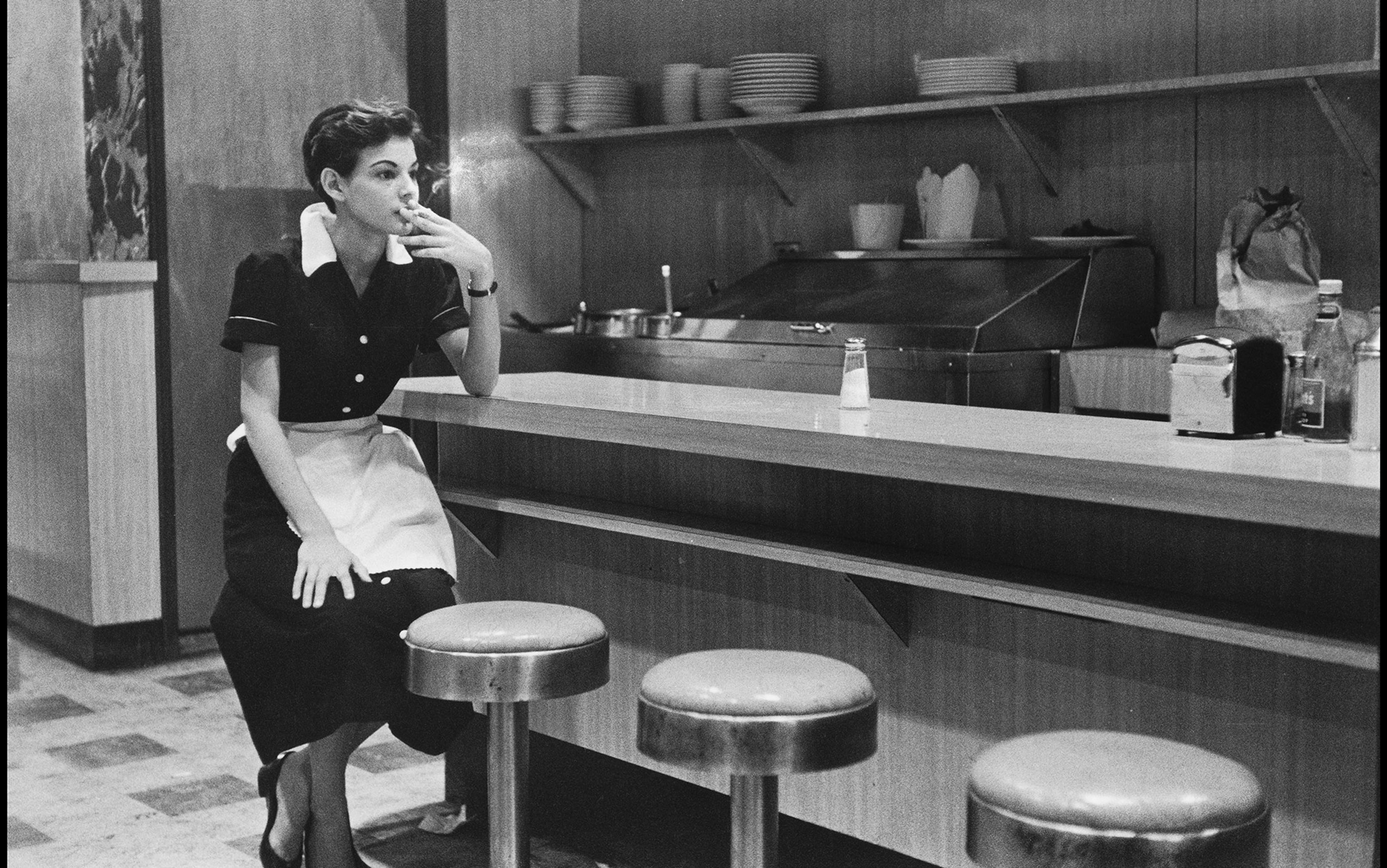 Vintage photo of a waitress in a diner sitting on a counter stool, smoking a cigarette, with kitchen equipment in the background.