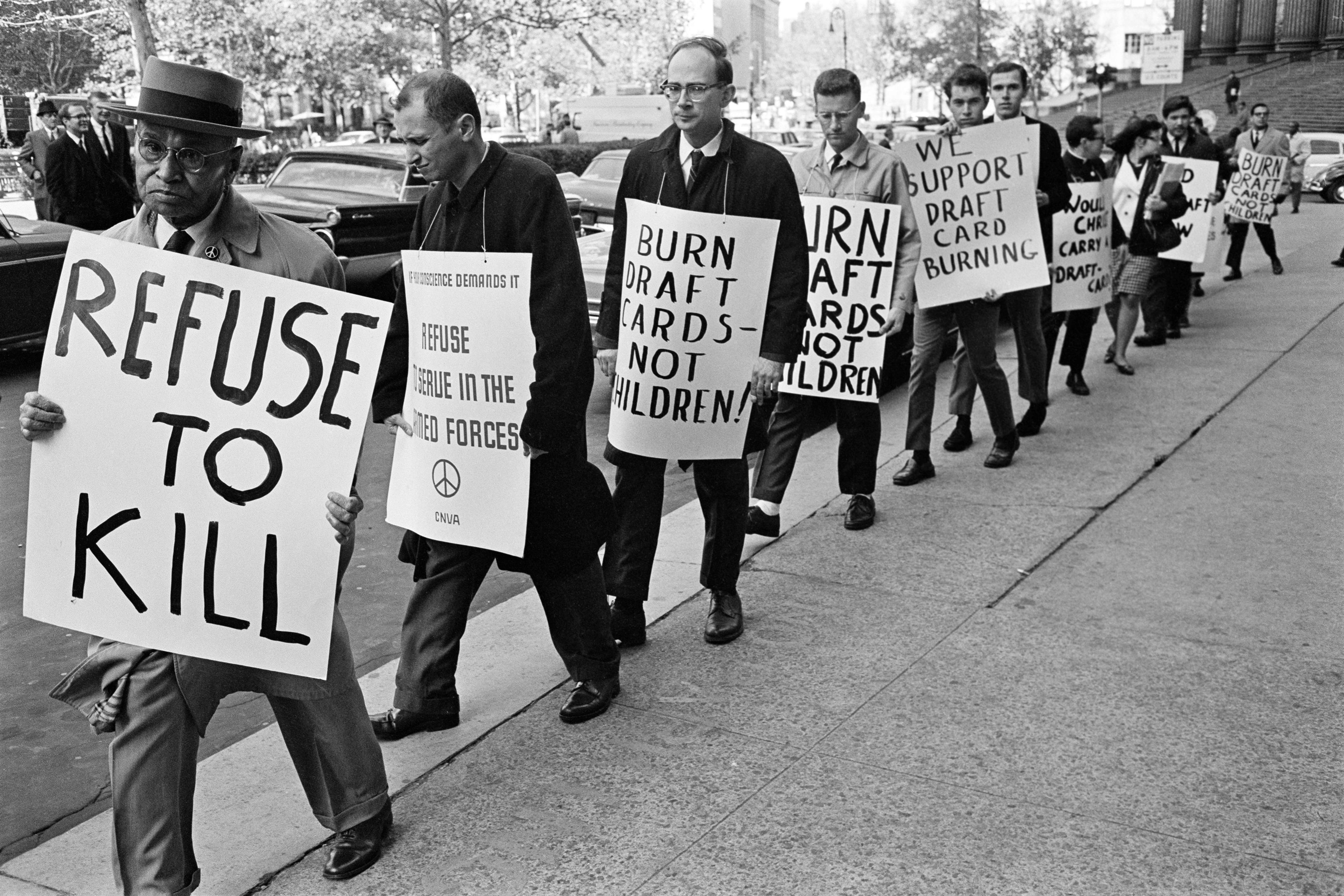 Black and white photo of a line of people marching on a city street holding signs protesting against the draft for the Vietnam War.