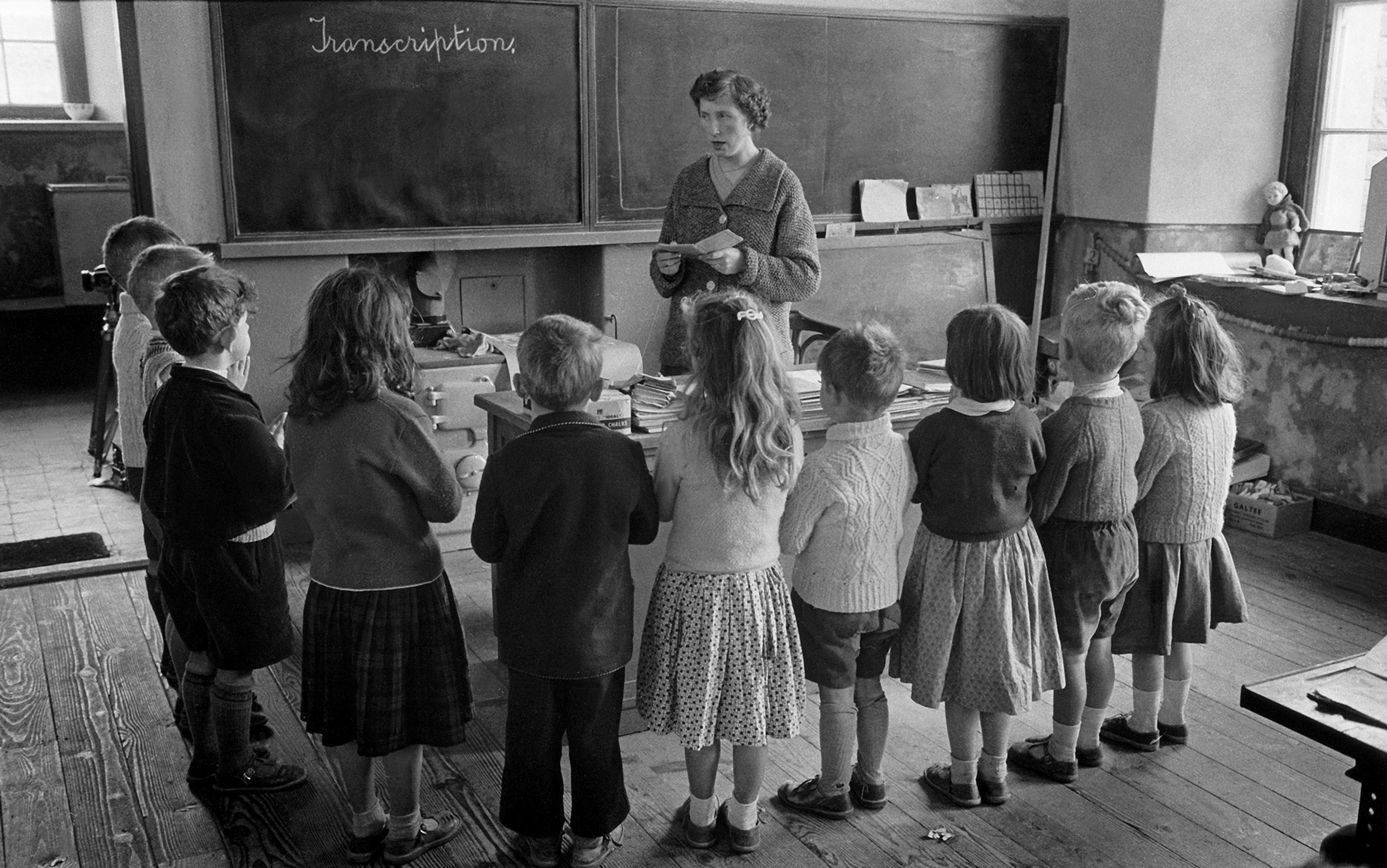 Children stand in a line facing a teacher in a classroom, with “Transcription” written on the blackboard behind her.