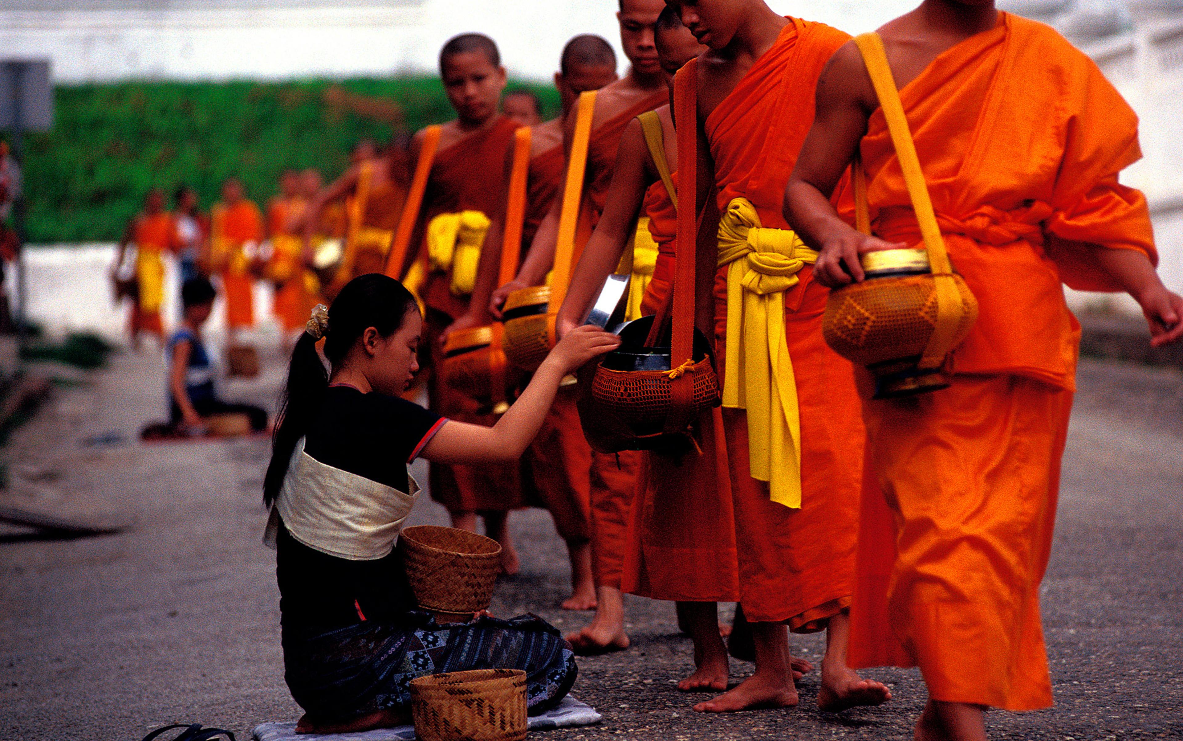 A woman offering alms to a line of monks in orange robes holding bowls in a traditional ceremony on a street.