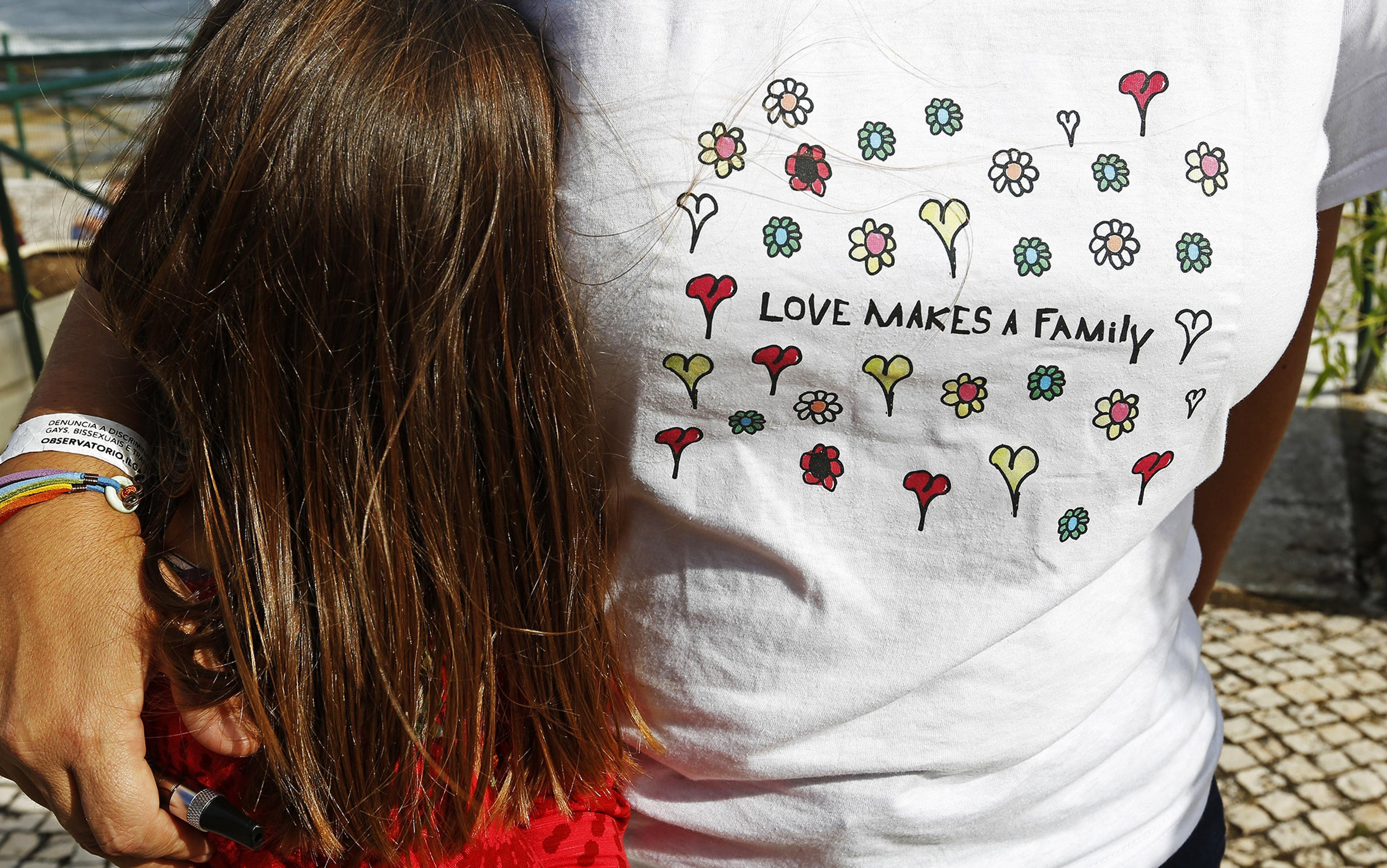 A person wearing a shirt with flowers and the text “Love Makes a Family” hugging a child, bracelet in rainbow colours.
