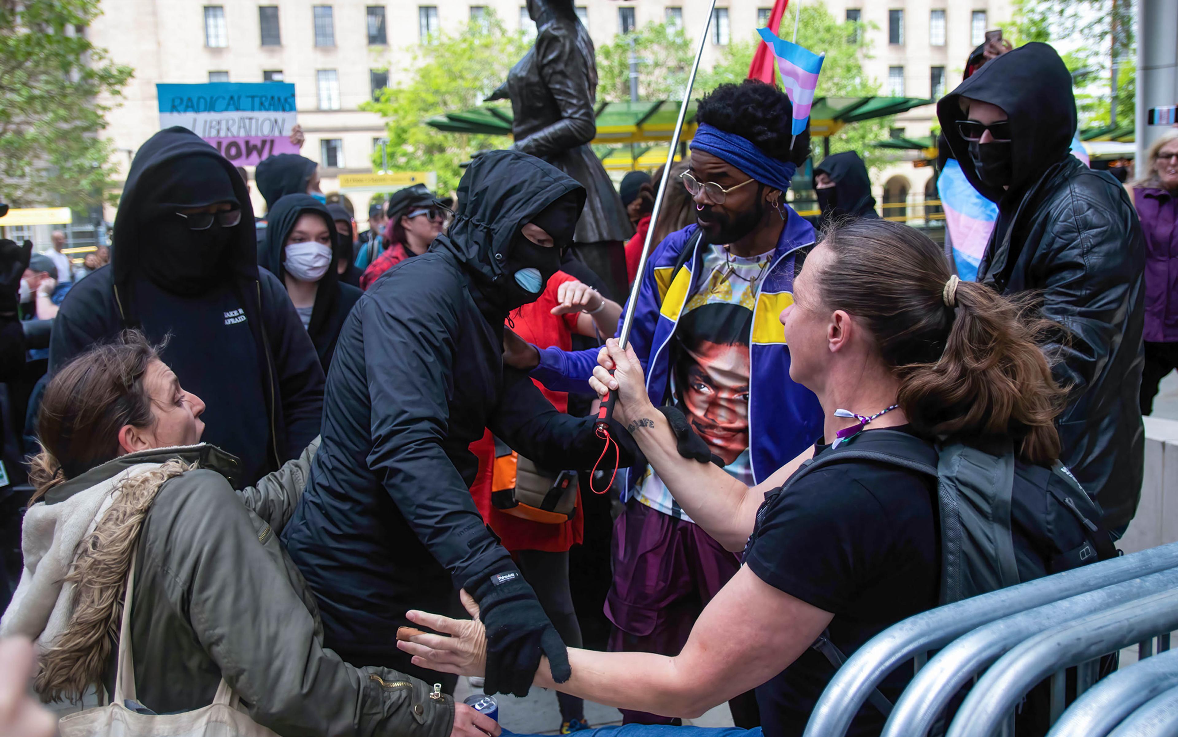 People in masks and hoods interacting in a crowd, some holding flags and signs, near a metal barricade in an outdoor urban setting.