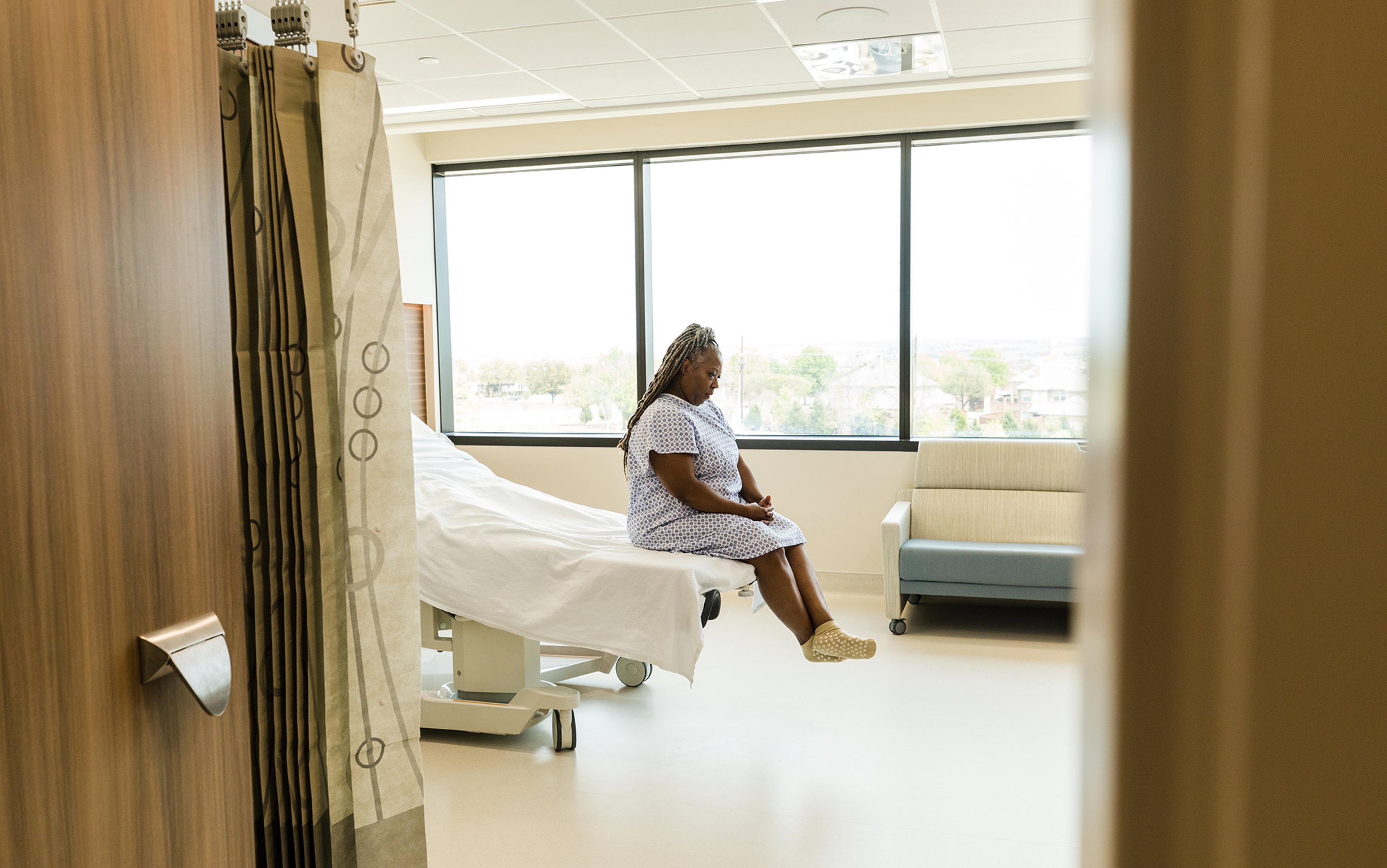 A person in a hospital gown sitting on a bed in a bright patient room, large window and privacy curtain visible.