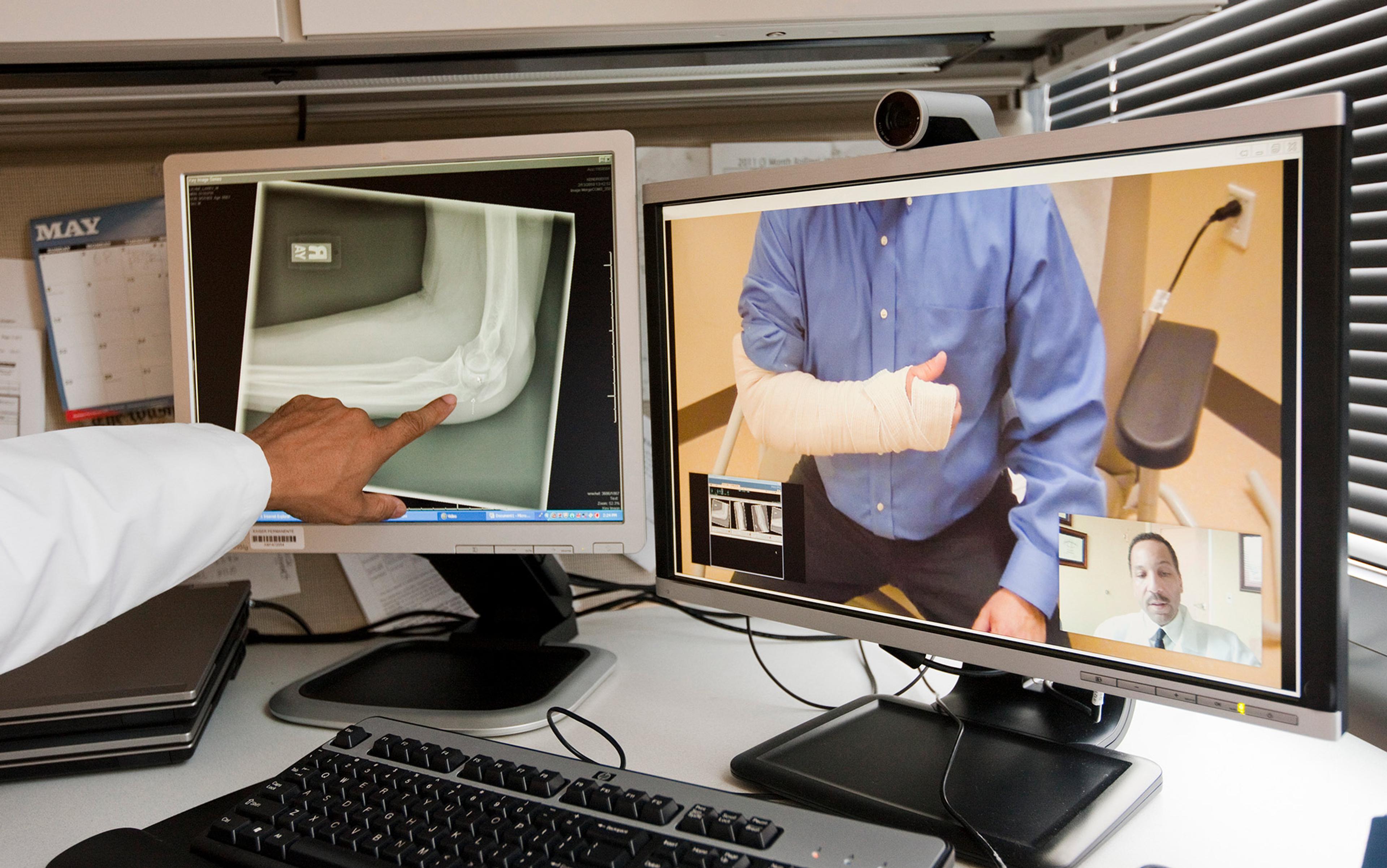A doctor points at an X-ray on a monitor while conducting a video consultation with a patient who has a bandaged arm, shown on a second monitor.