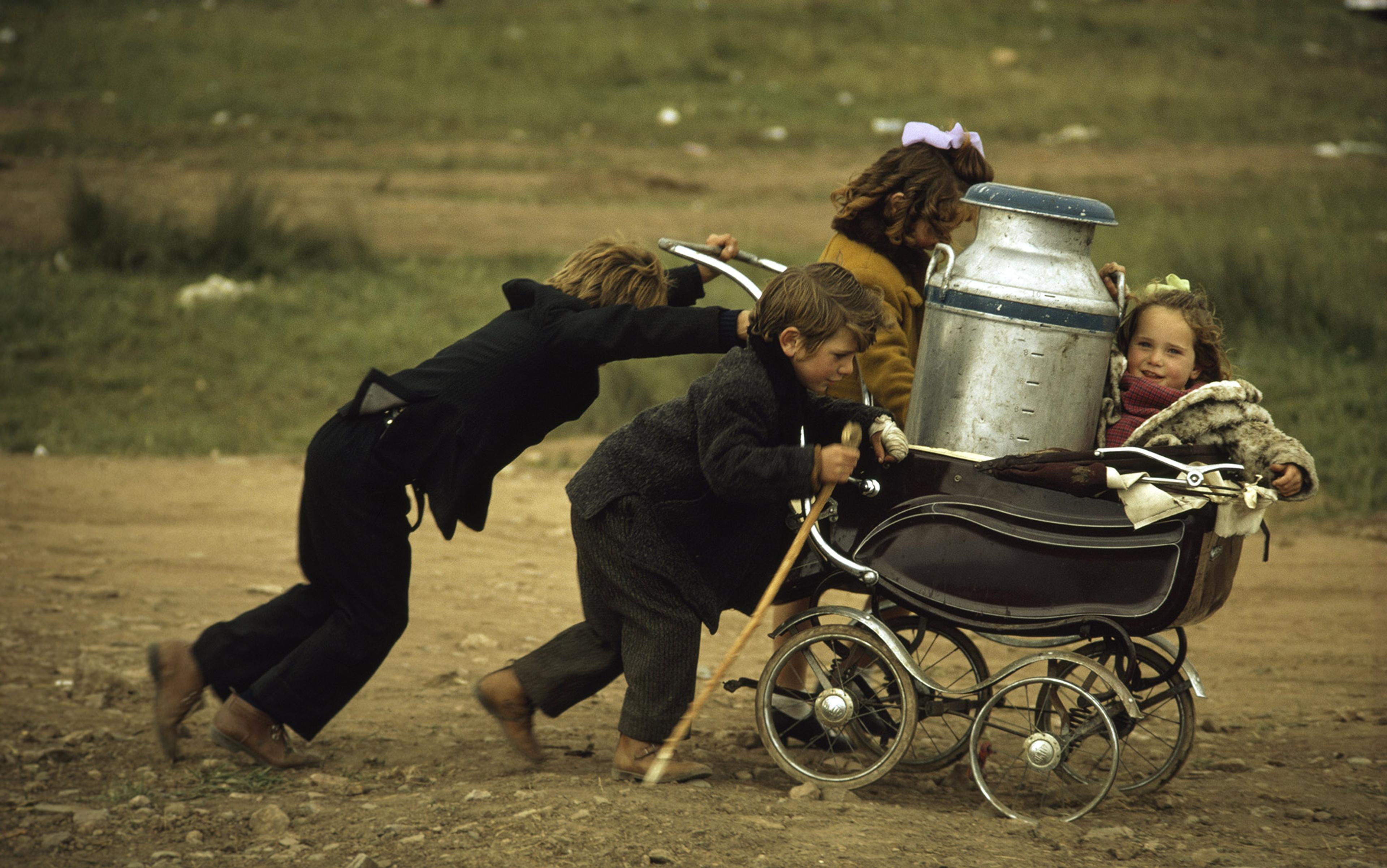 Three children pushing a pram holding another child and a milk churn on a dirt path, with grass in the background.