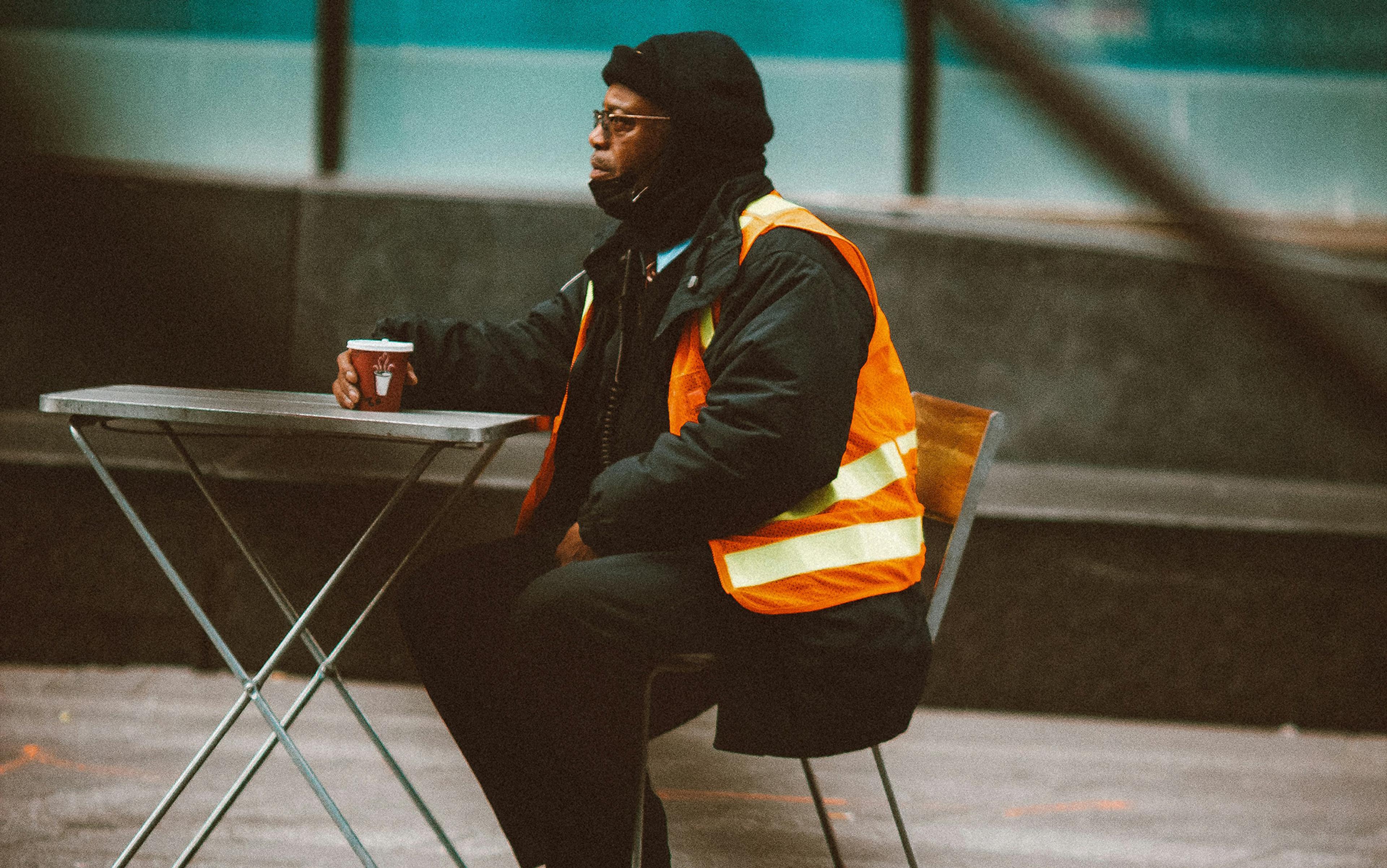 A person in a hi-vis vest sitting outdoors at a table with a drink, looking into the distance.
