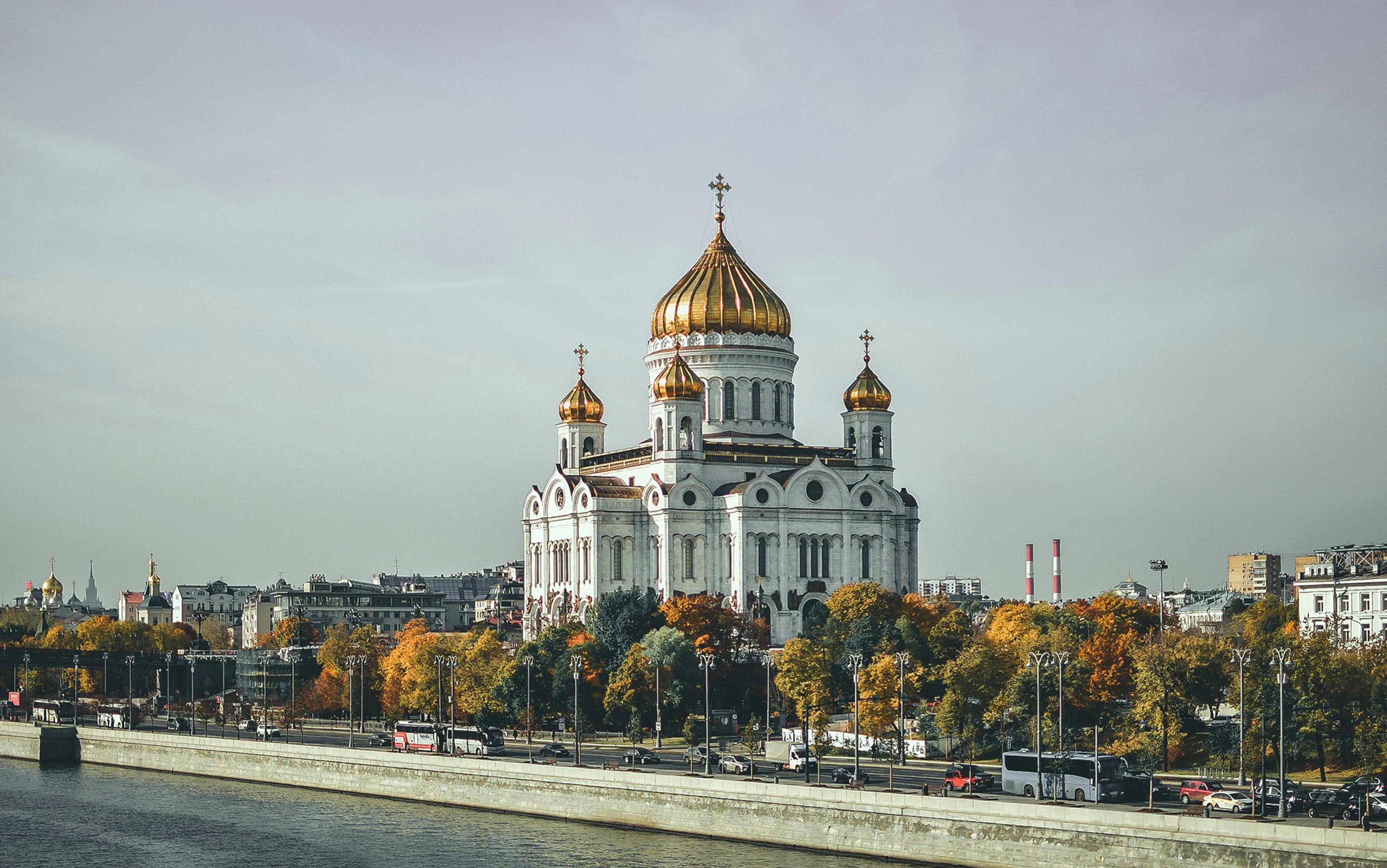 A large white cathedral with golden domes near a river, surrounded by trees with autumn colours.