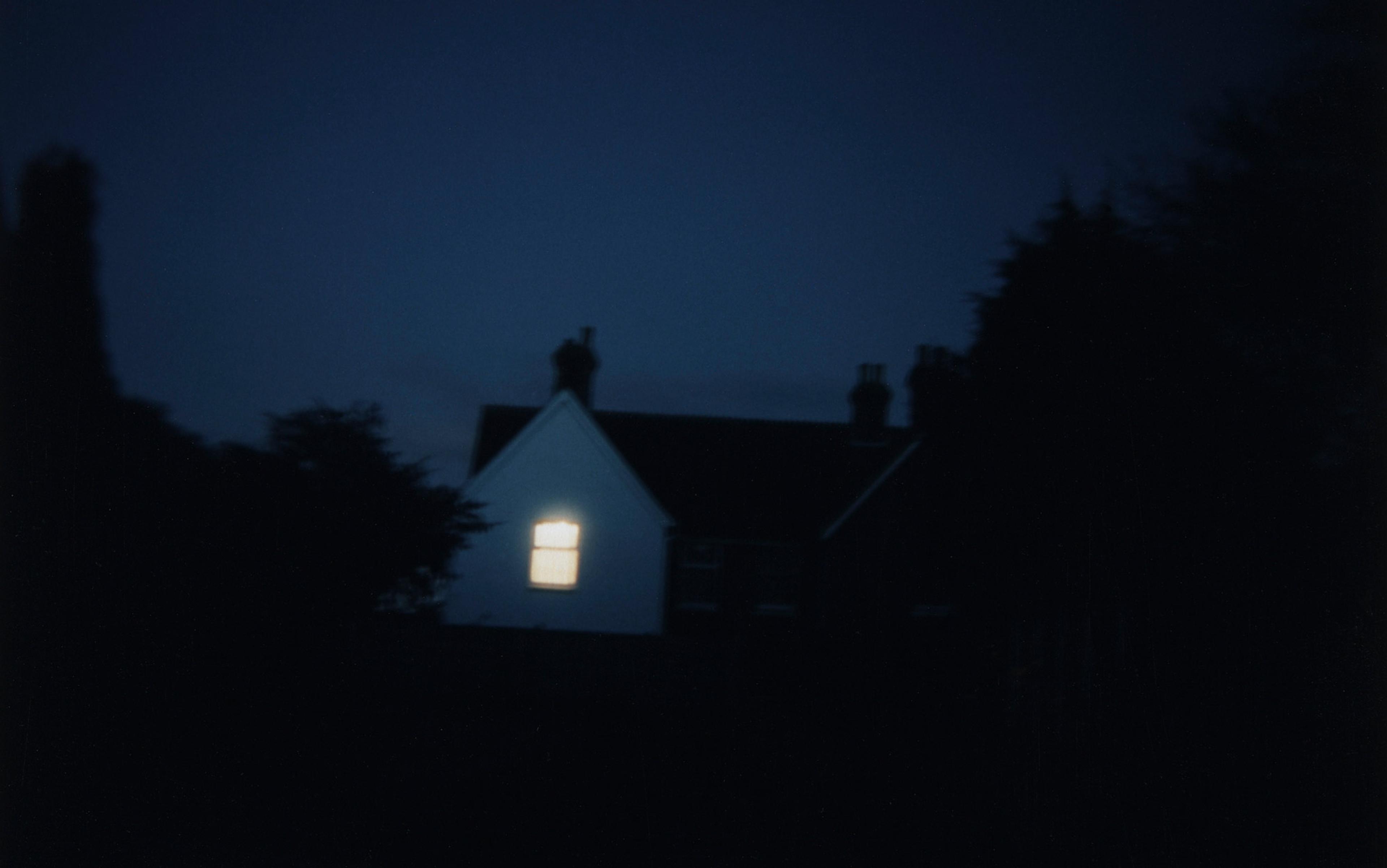 A house at night with its single window lit, surrounded by dark shadows and trees against a deep blue sky.