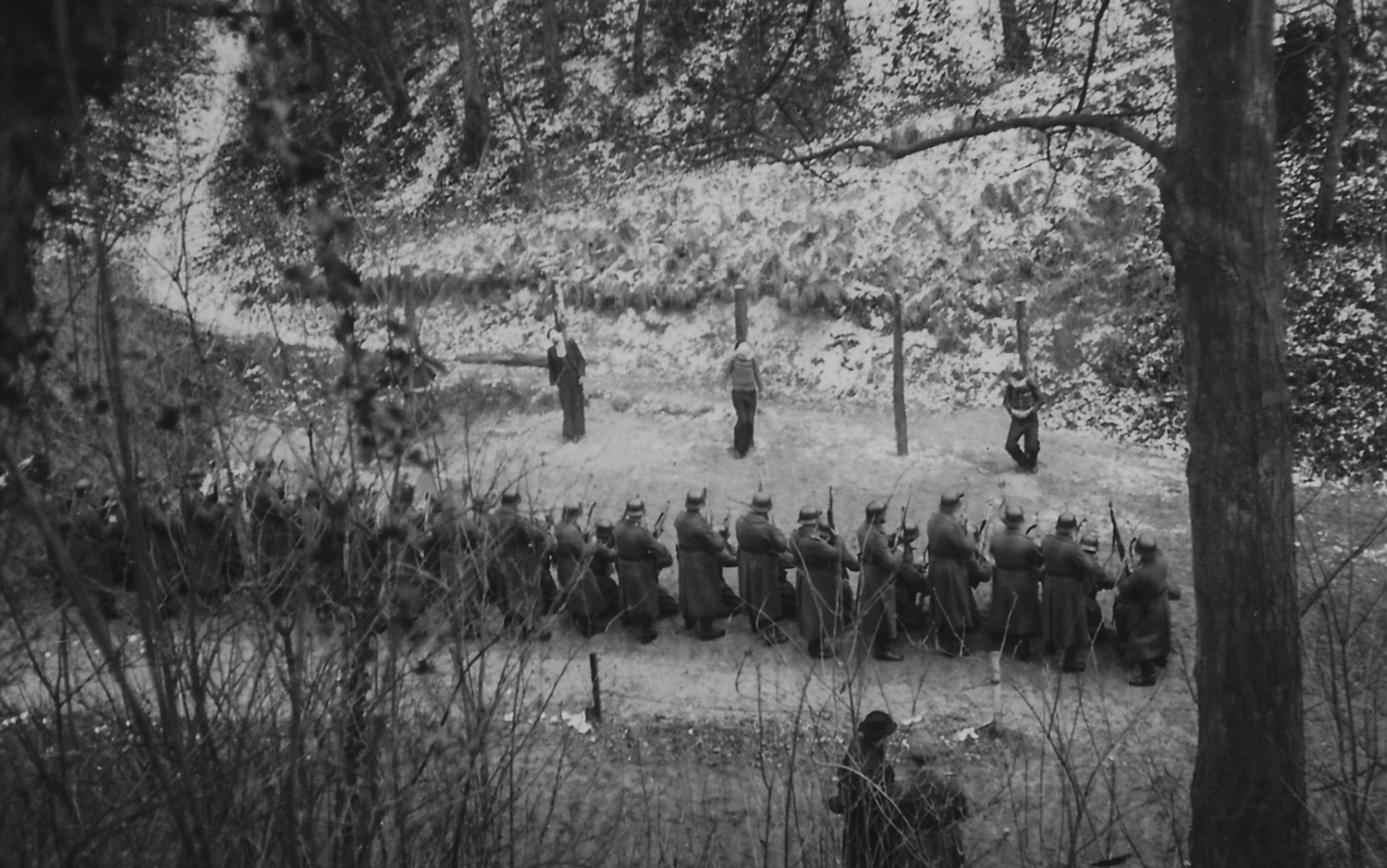 Black and white photo of a firing squad in a wooded area with snow on the ground, soldiers aiming rifles at standing figures.