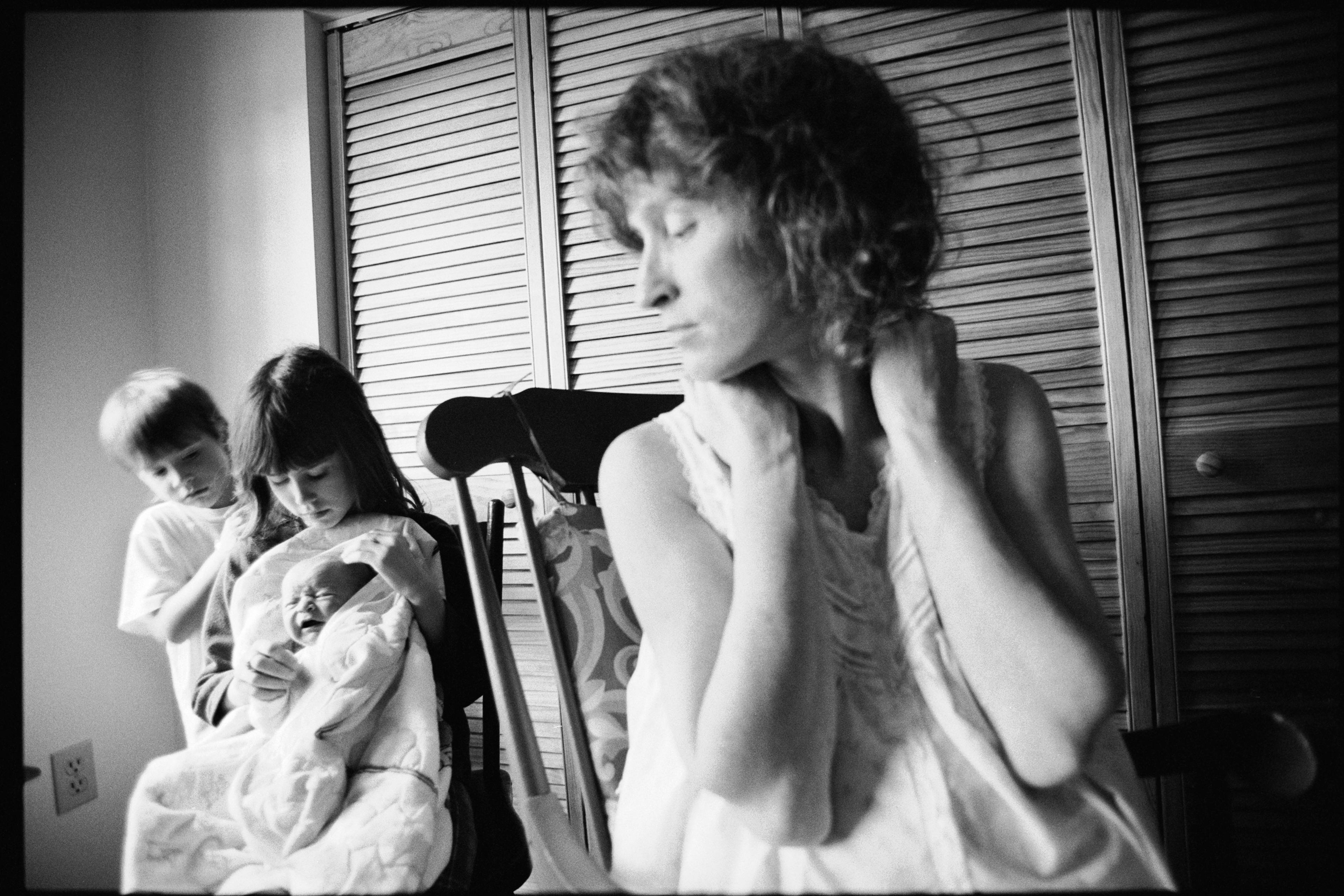 Black and white photo of a thoughtful woman and two children, one holding a crying baby, indoors near wooden doors.