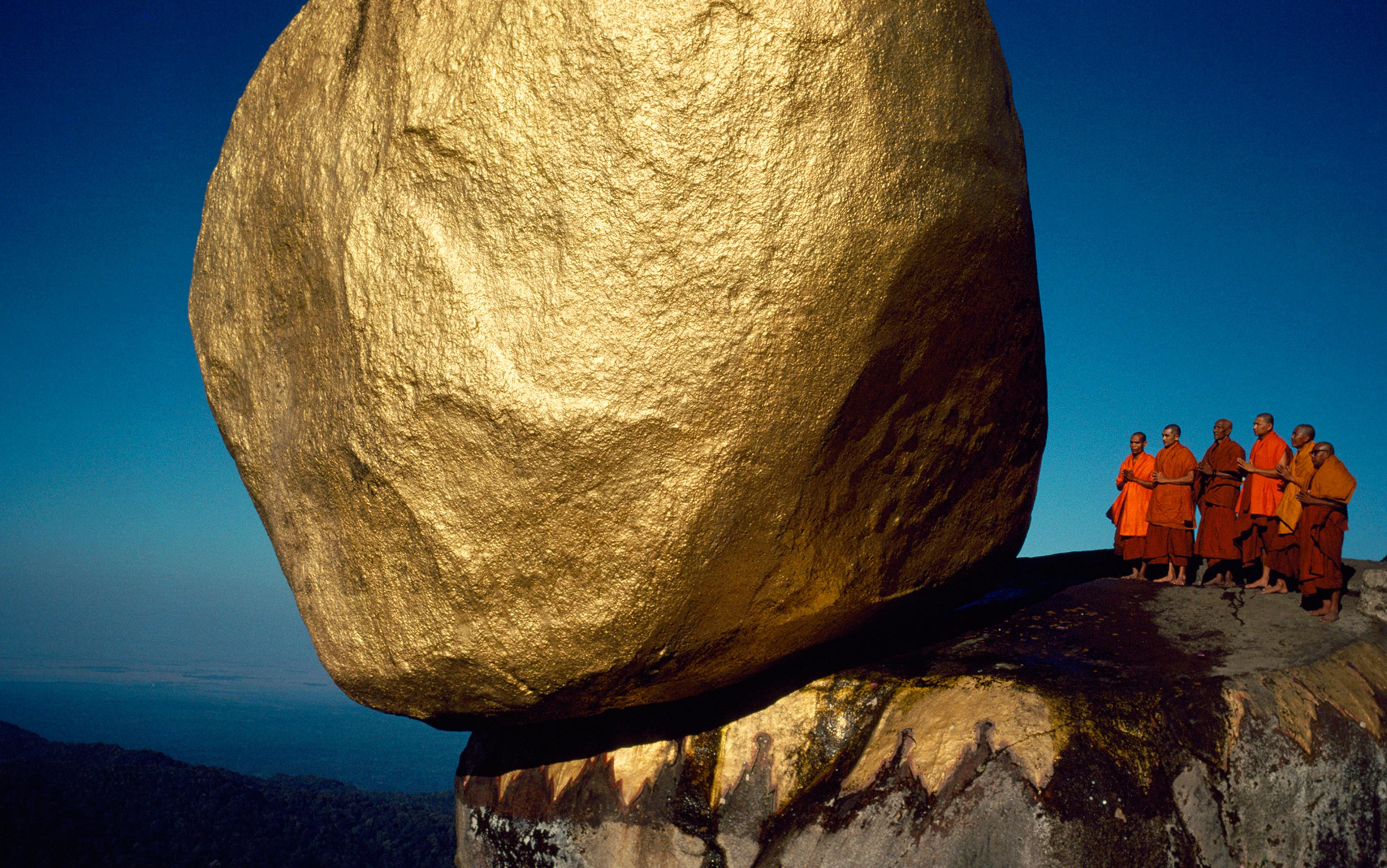 Monks in orange robes standing near the Golden Rock in Myanmar, with a bright blue sky in the background.