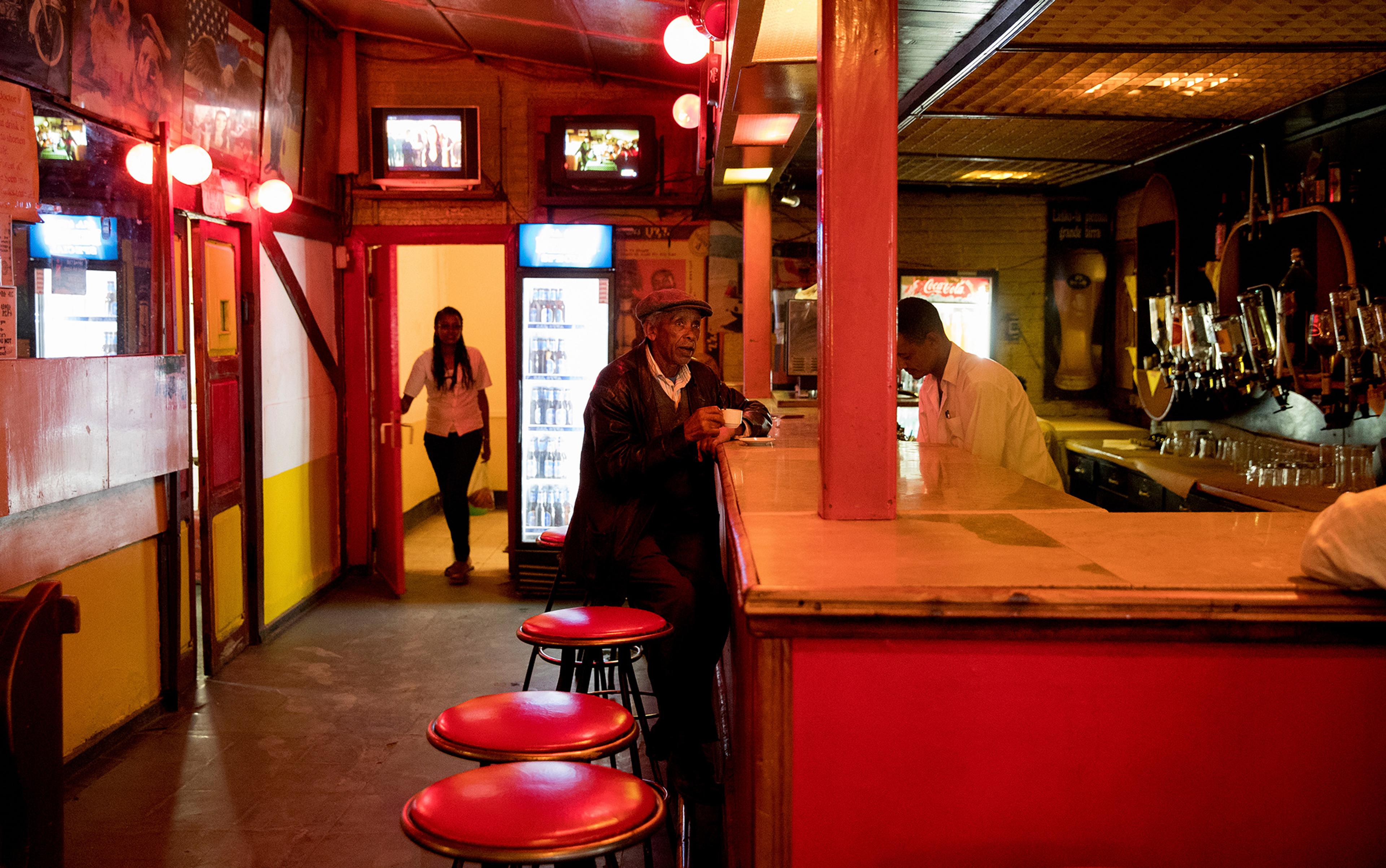 A dimly lit bar with red decor, a man sitting at the counter, a bartender, and a woman entering through a door.