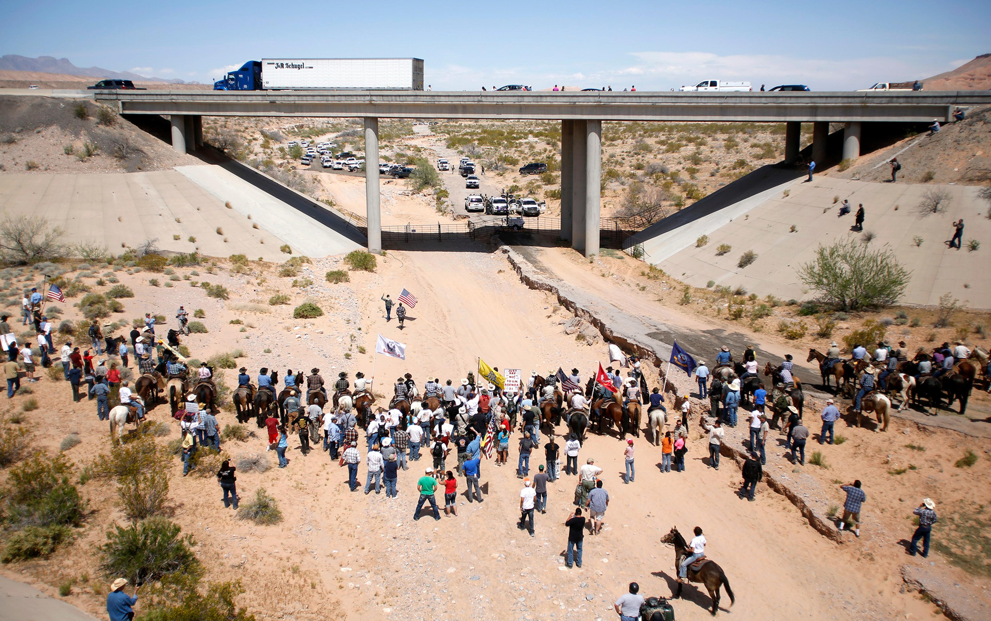 A group of people, some on horseback, gather in a desert area under a motorway bridge. Some have flags and banners. Police cars can be seen in the background.