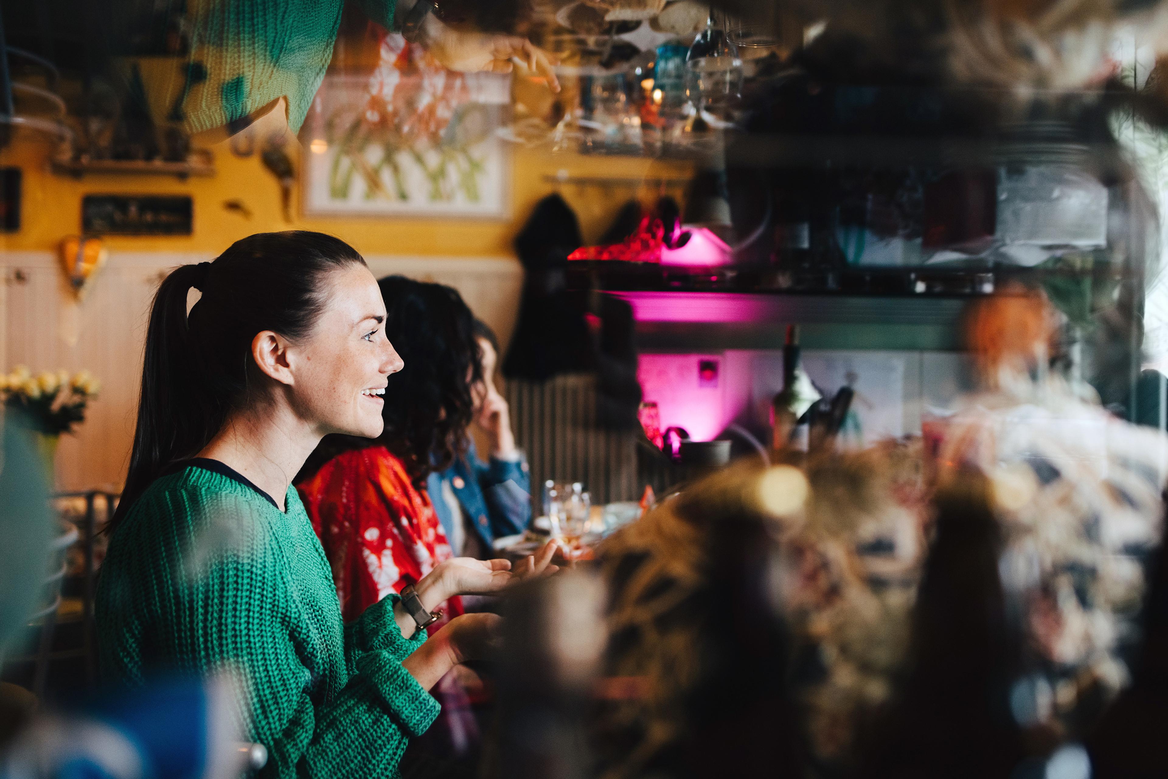 Photo of a smiling woman in a green sweater talking with friends in a cosy, colourful cafe interior.