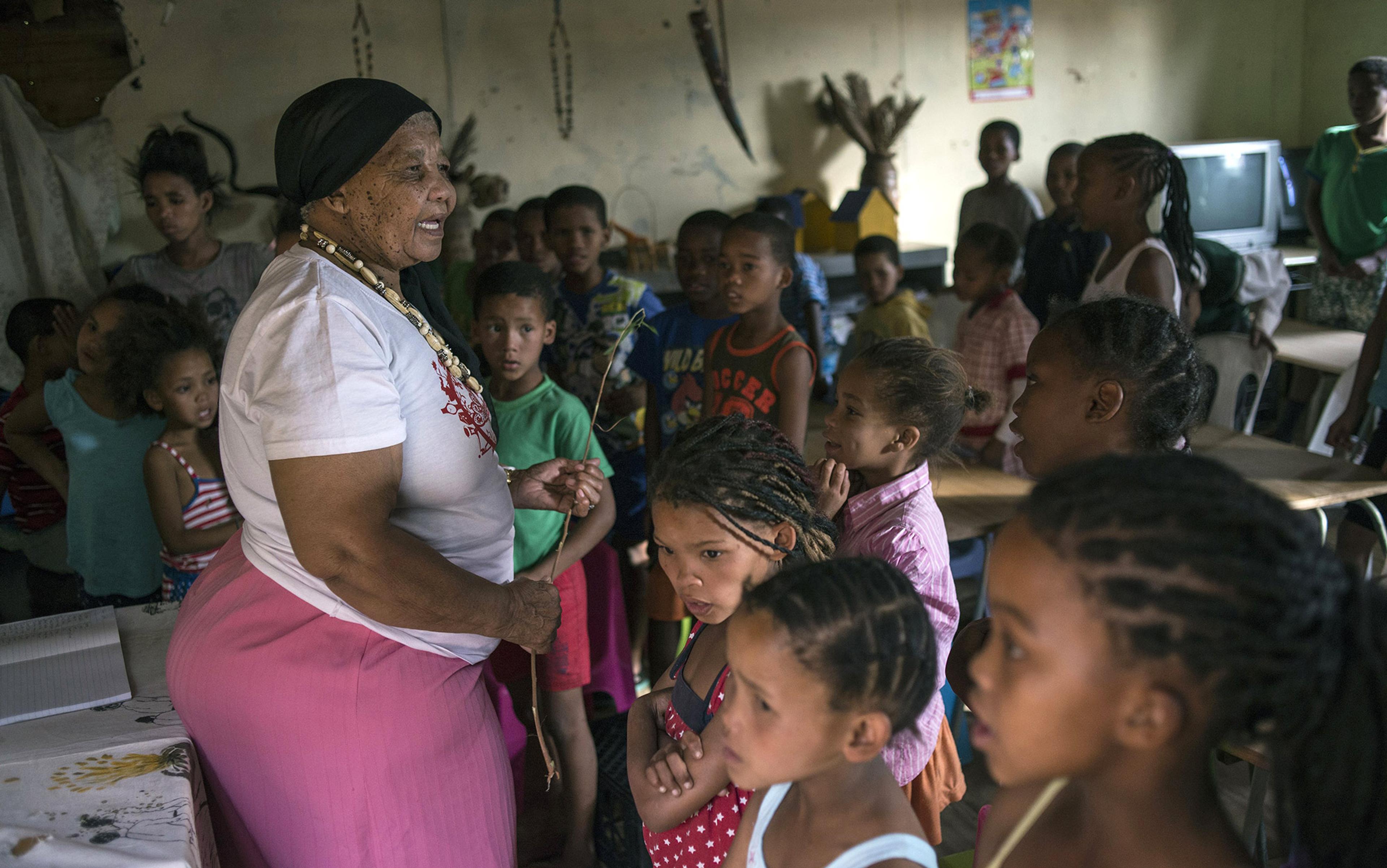 A woman in a pink skirt and black headscarf interacting with a group of children in a classroom setting.