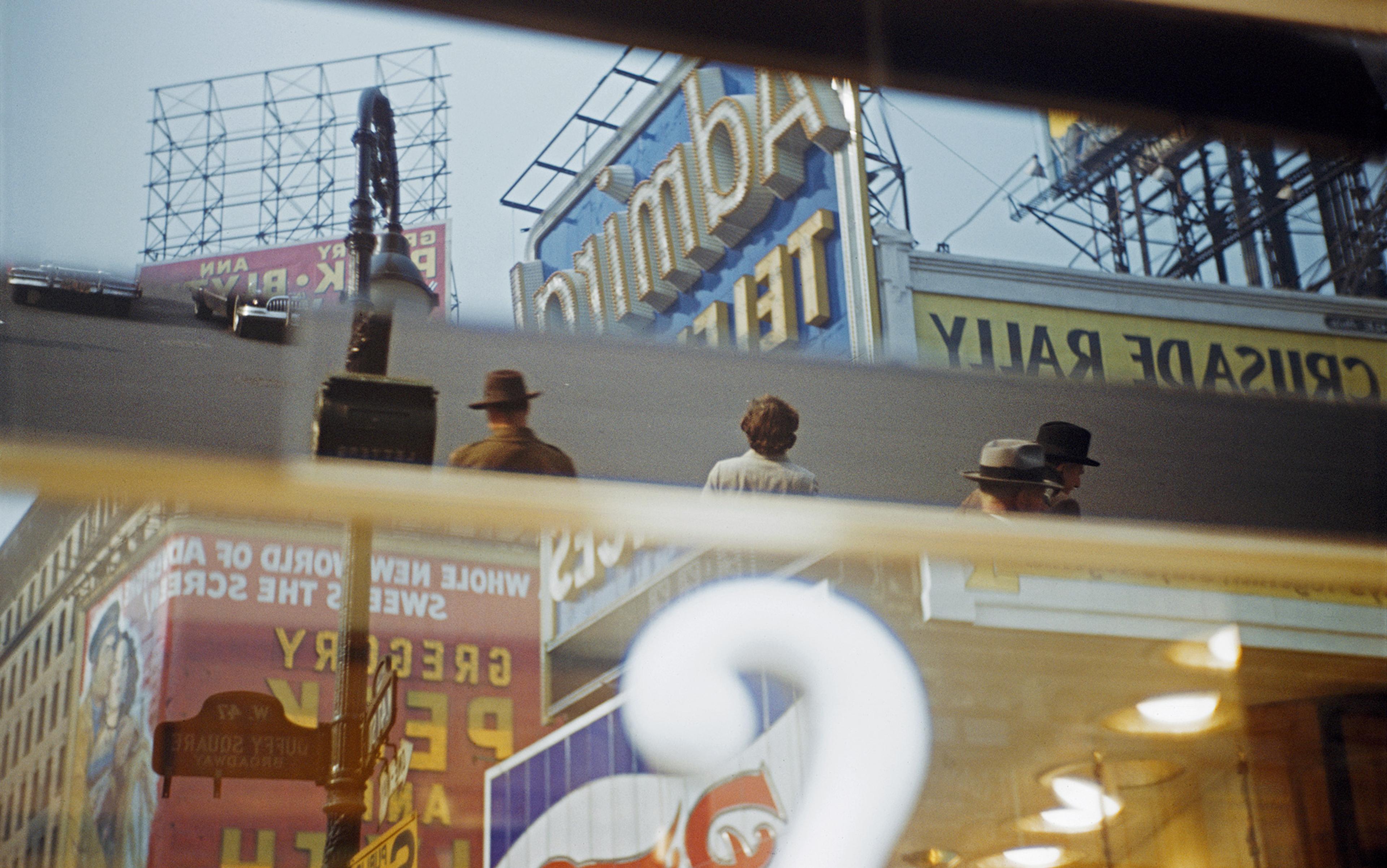 Reflection of an urban street scene, including people wearing hats and billboards, on a window with a white question mark on the glass.