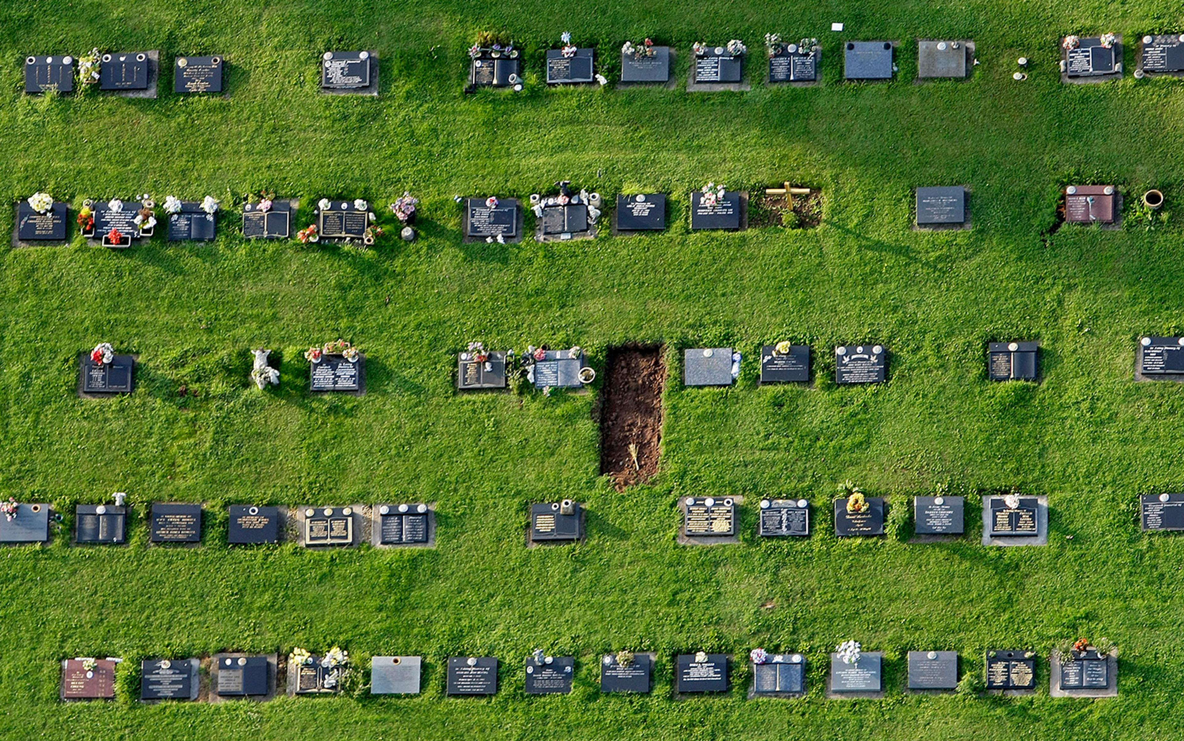 Aerial view of a cemetery with rows of headstones on grass, featuring a freshly dug grave in the middle right section.