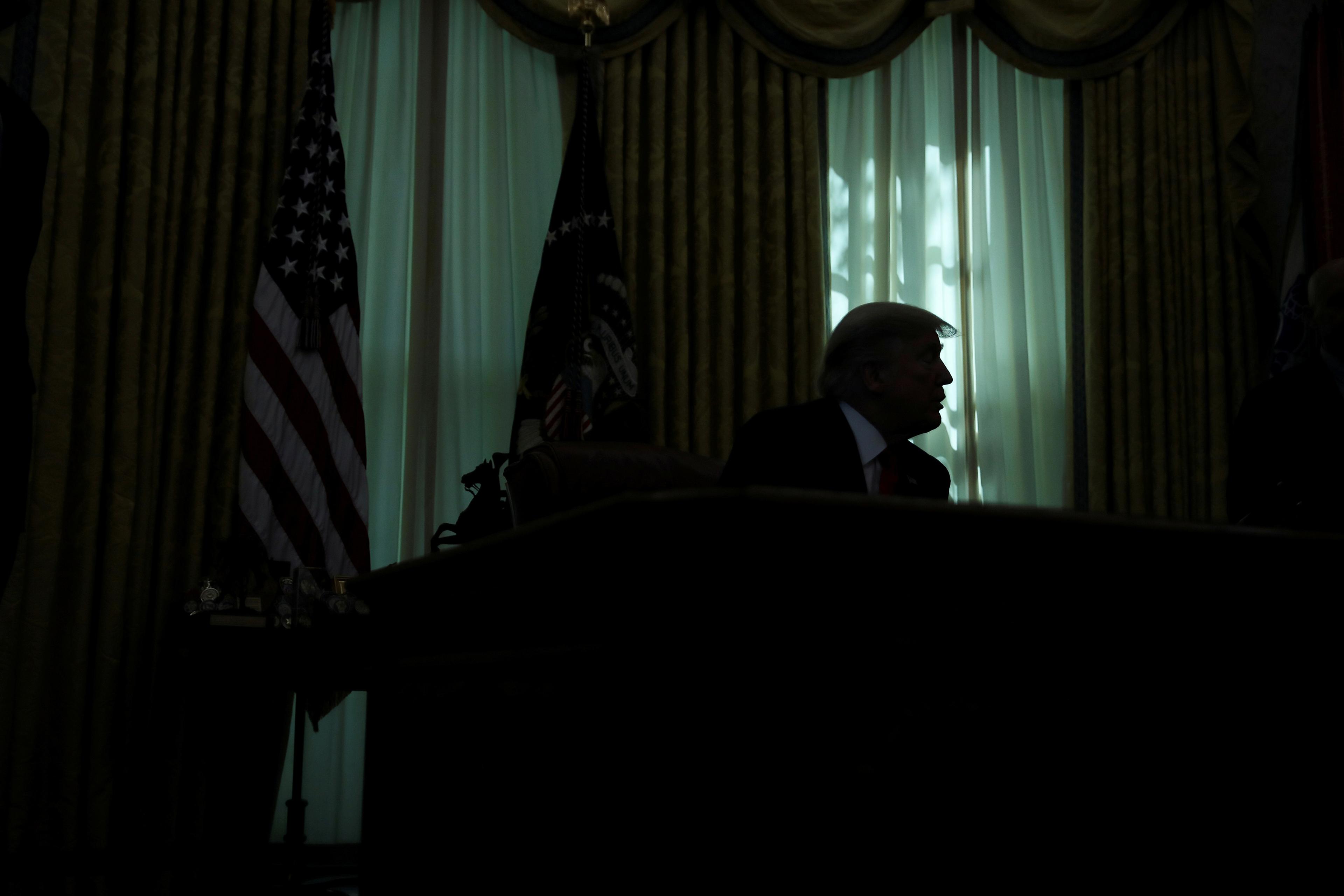 Silhouetted figure at a desk with US flag nearby, backlit by sunlight through curtains.