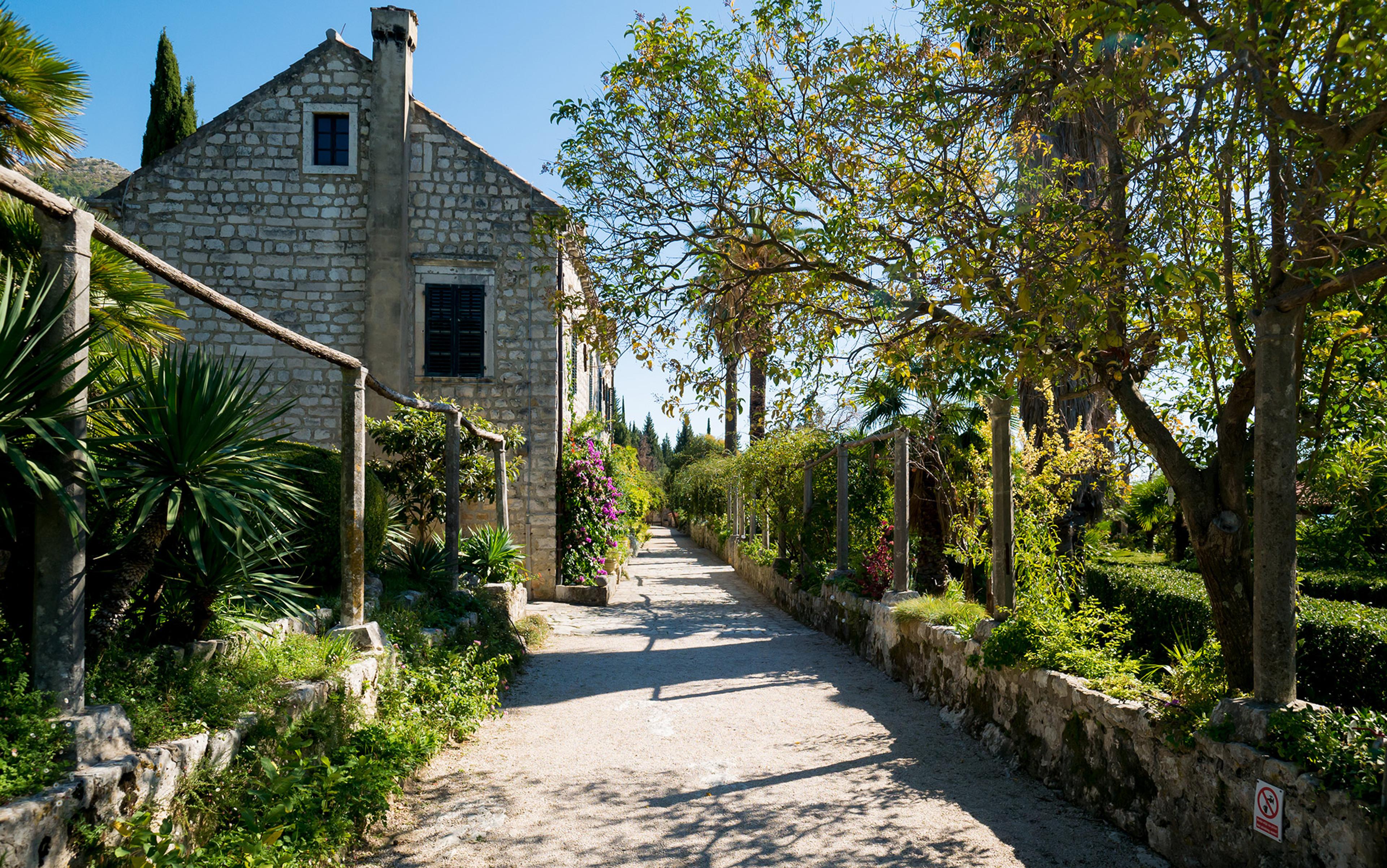Photo of a rustic stone house with a shaded garden path lined with lush greenery and flowering plants under a clear blue sky.