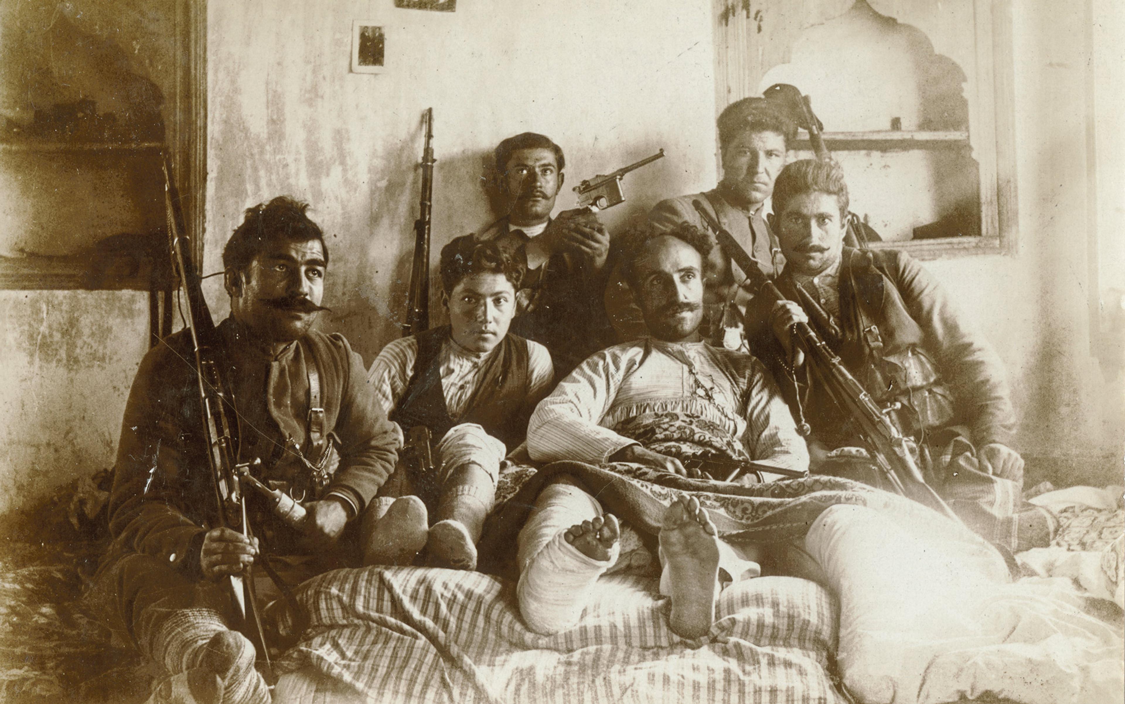 Vintage photo of six men holding rifles and a handgun on a bed in a room with textured walls; the central figure is lying down with on leg in bandages.