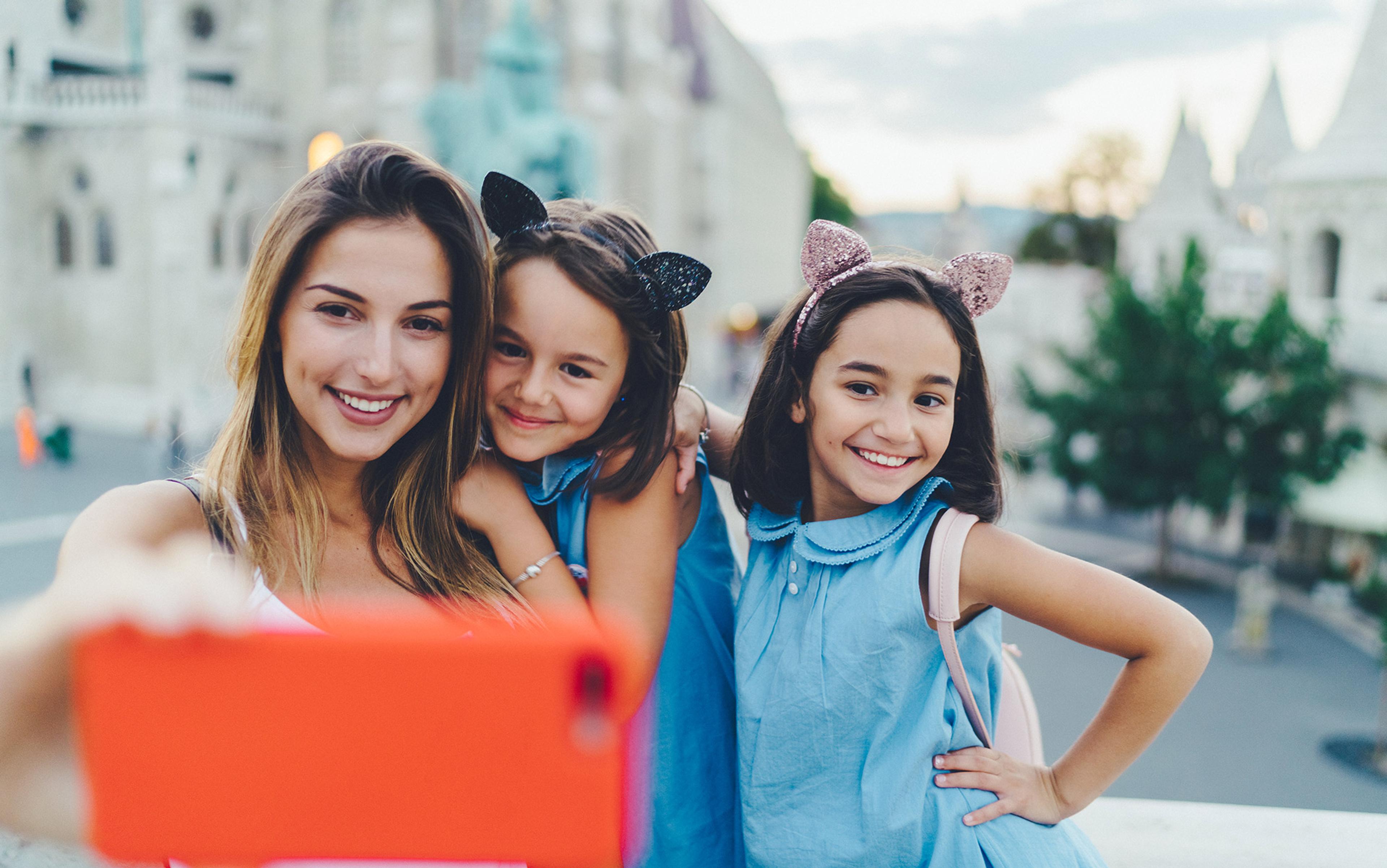 A woman and two girls in blue dresses with cat-ear headbands, smiling and taking a selfie outdoors.