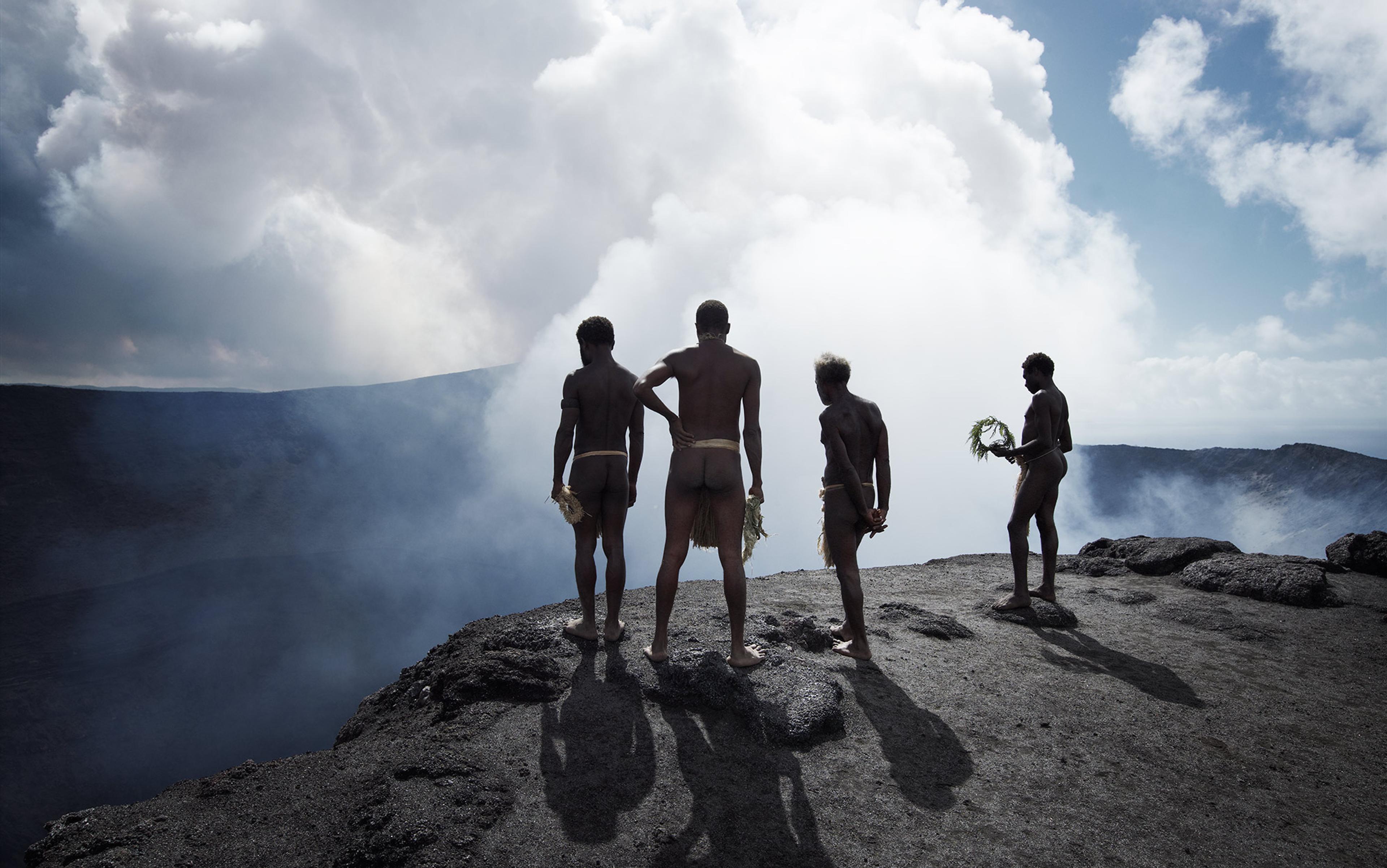 Four people standing on a rocky cliff edge surrounded by mist and clouds, wearing minimal traditional attire and holding foliage.