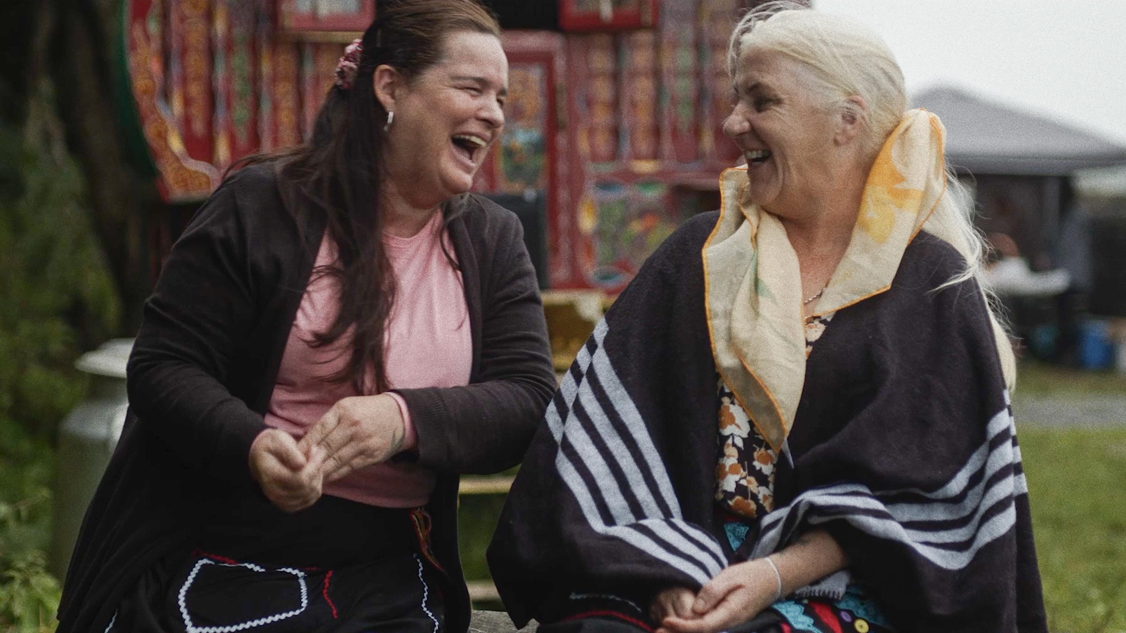 Photo of two women sitting outdoors laughing with a colourful backdrop in the background.