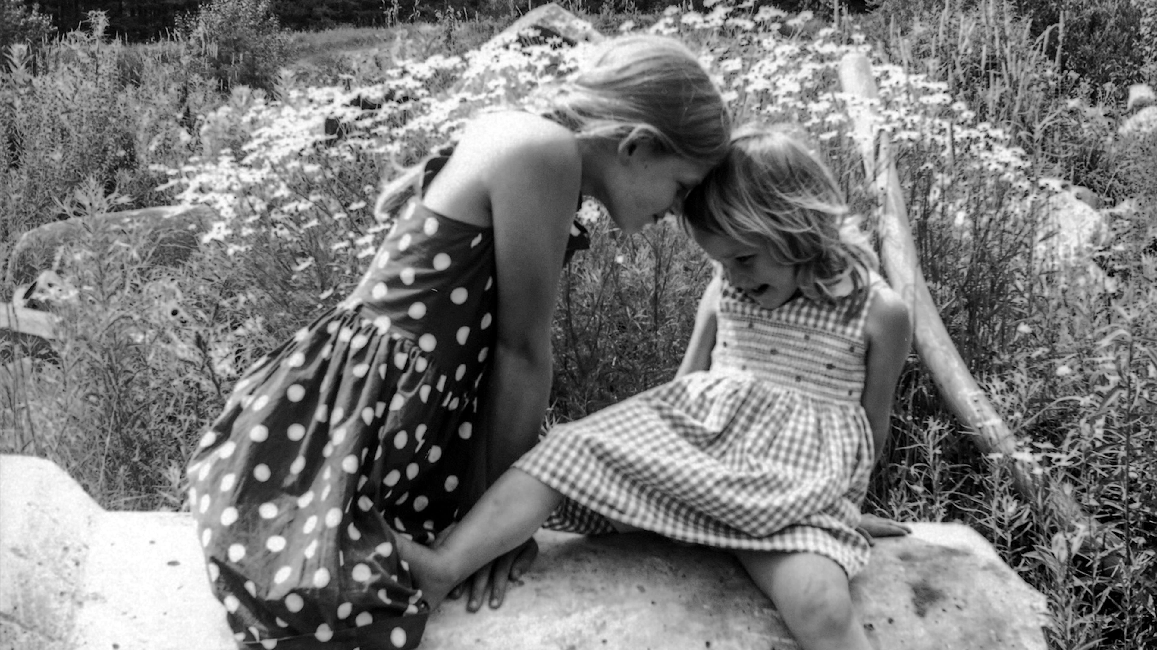 Black and white photo of two girls in dresses touching foreheads while sitting on a rock in a field of daisies.